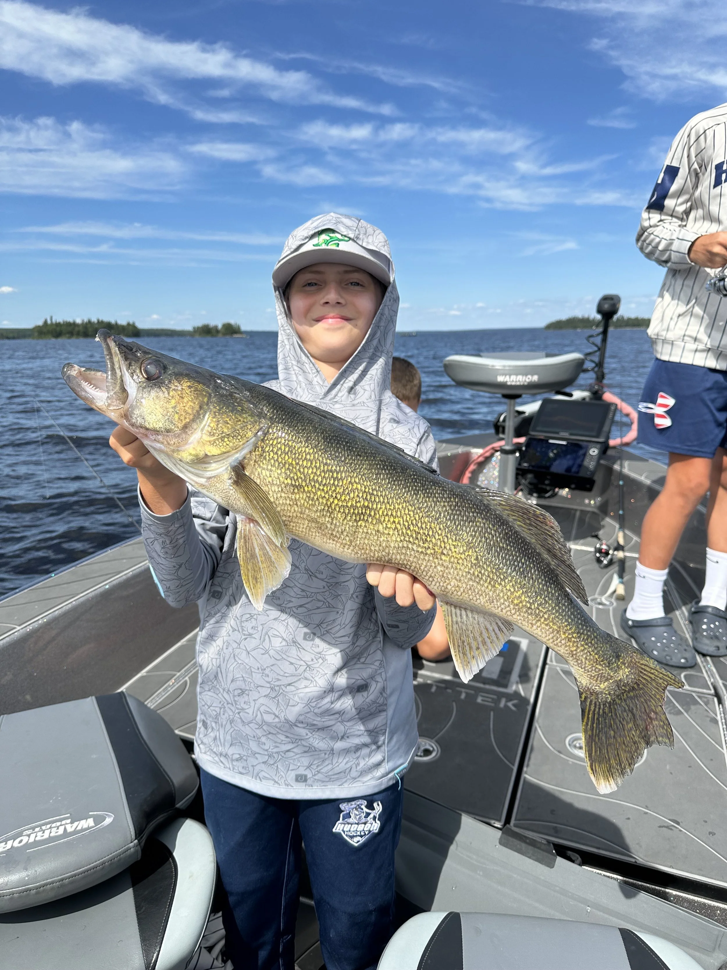 A boy on a boat holding a large fish he caught during a fishing trip on a lake under a partly cloudy sky.