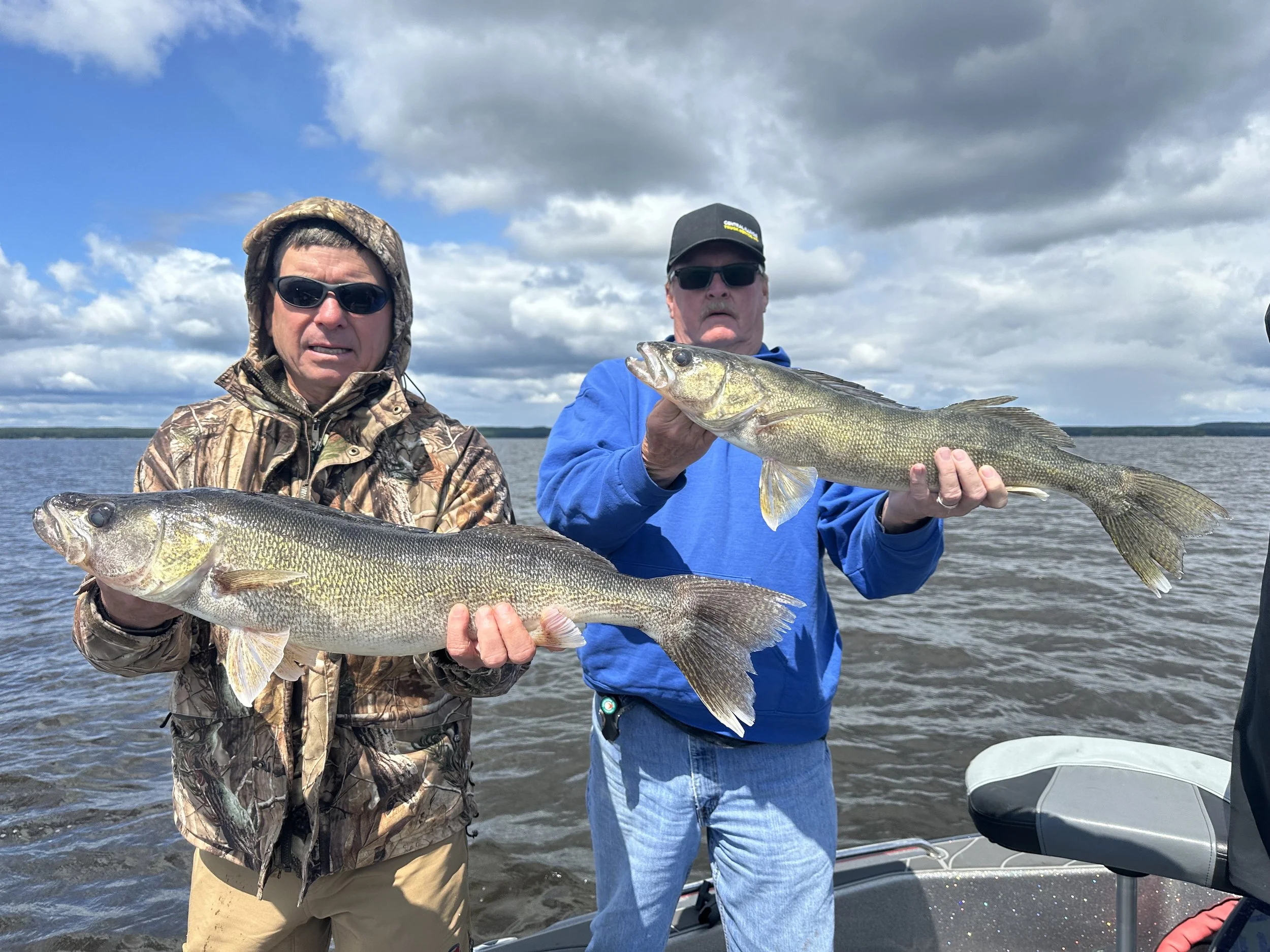 Two men on a boat holding large fish they caught during a fishing trip, with water and cloudy sky in the background.