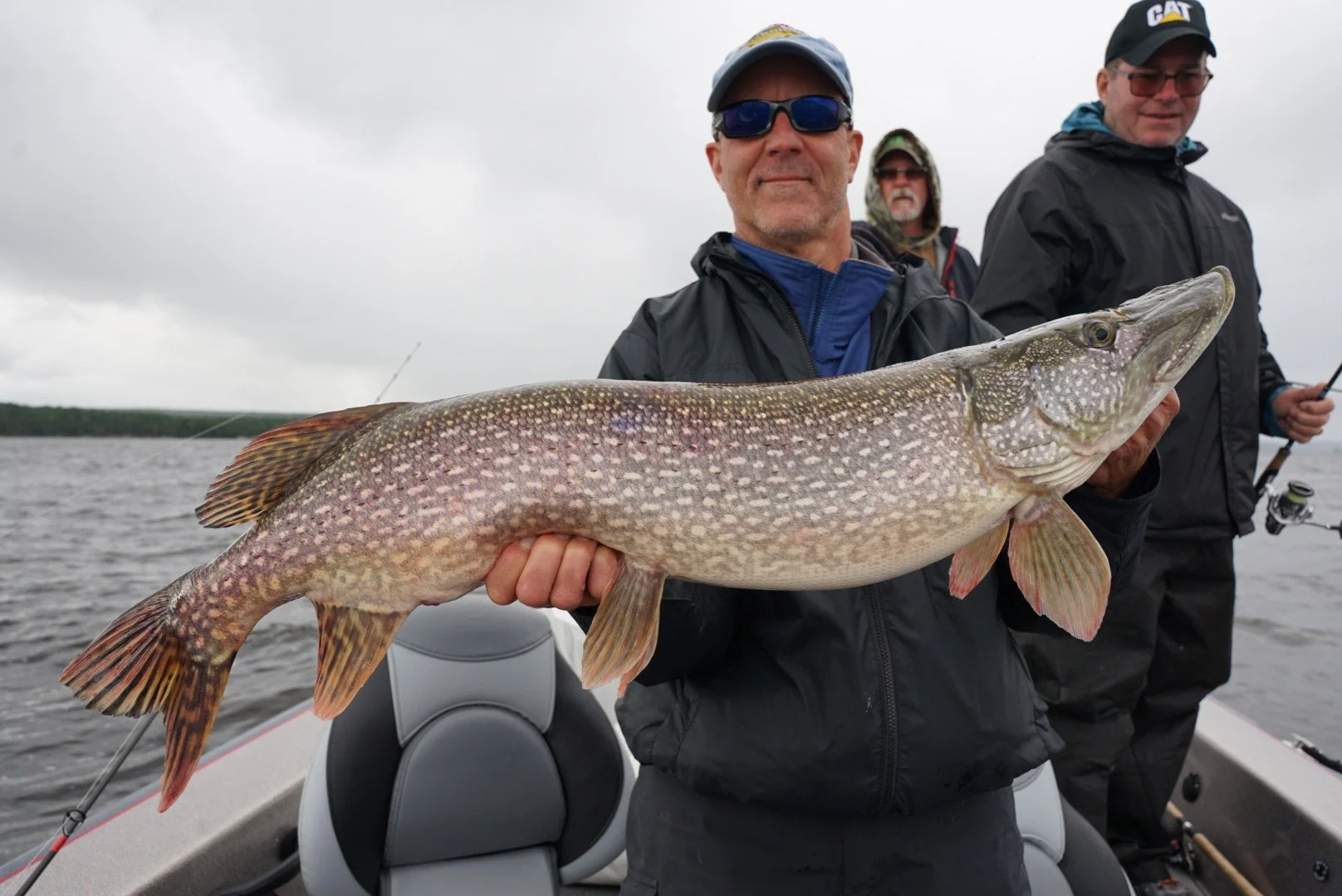 Man holding a large fish on a boat with two other men in the background, on a cloudy day.