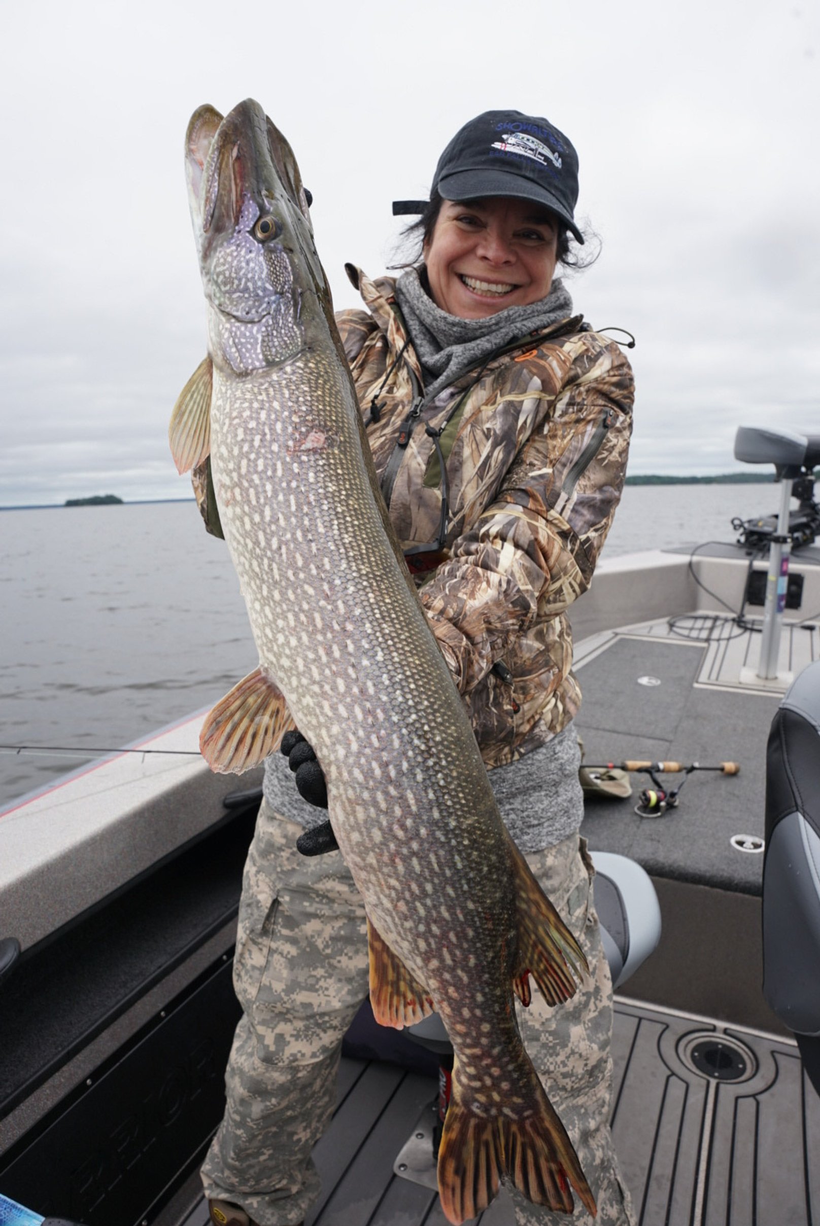 A woman in camouflage clothing and a black cap is smiling and holding a large fish on a boat in a waterbody, with fishing gear visible.