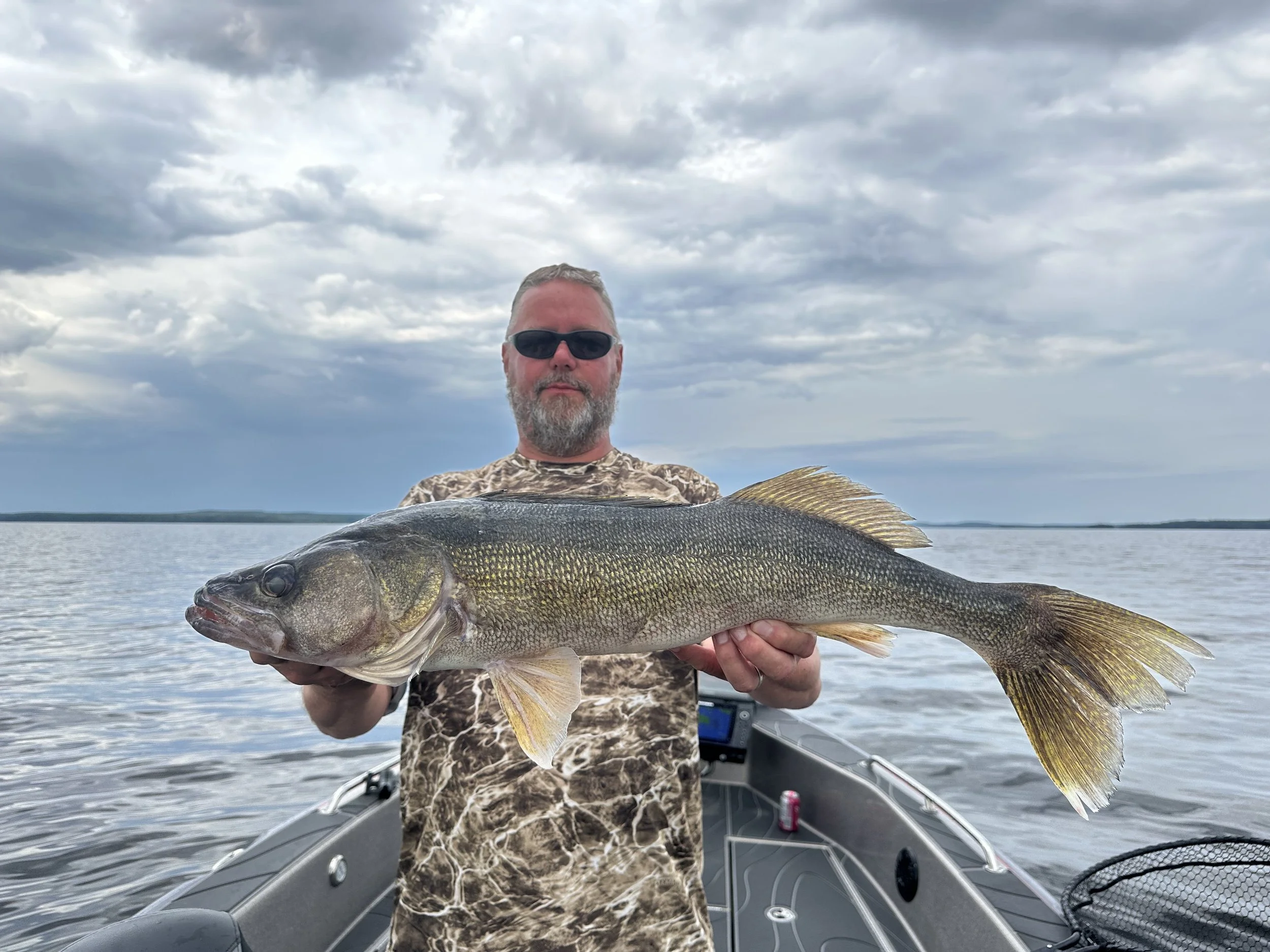A man with sunglasses and a beard holding a large fish on a boat in a lake with cloudy sky in the background.