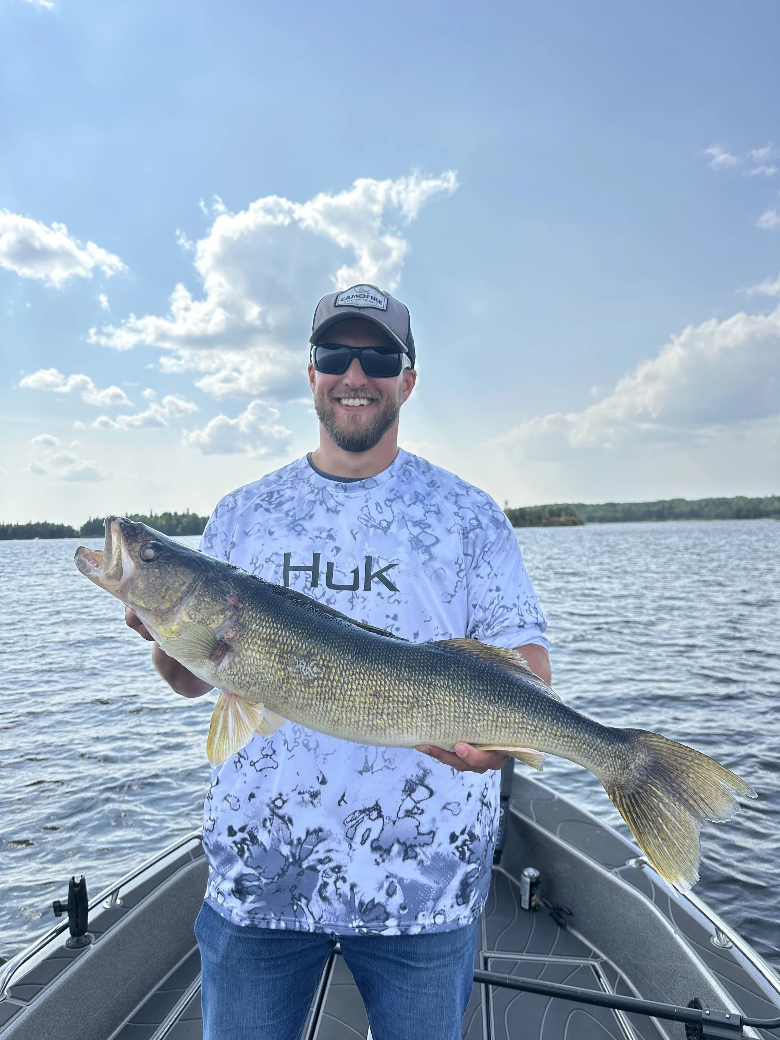 Man smiling on a boat holding a large fish in front of a lake with a cloudy sky.