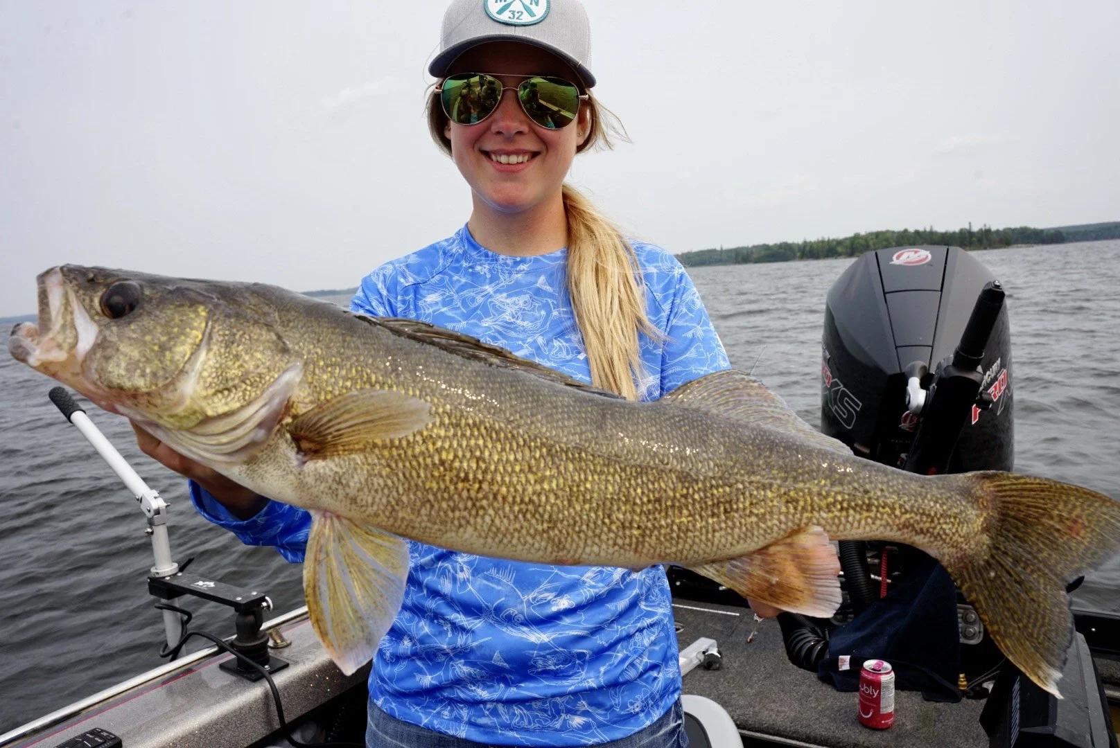 A woman in sunglasses and a baseball cap holding a large fish on a boat in a lake.