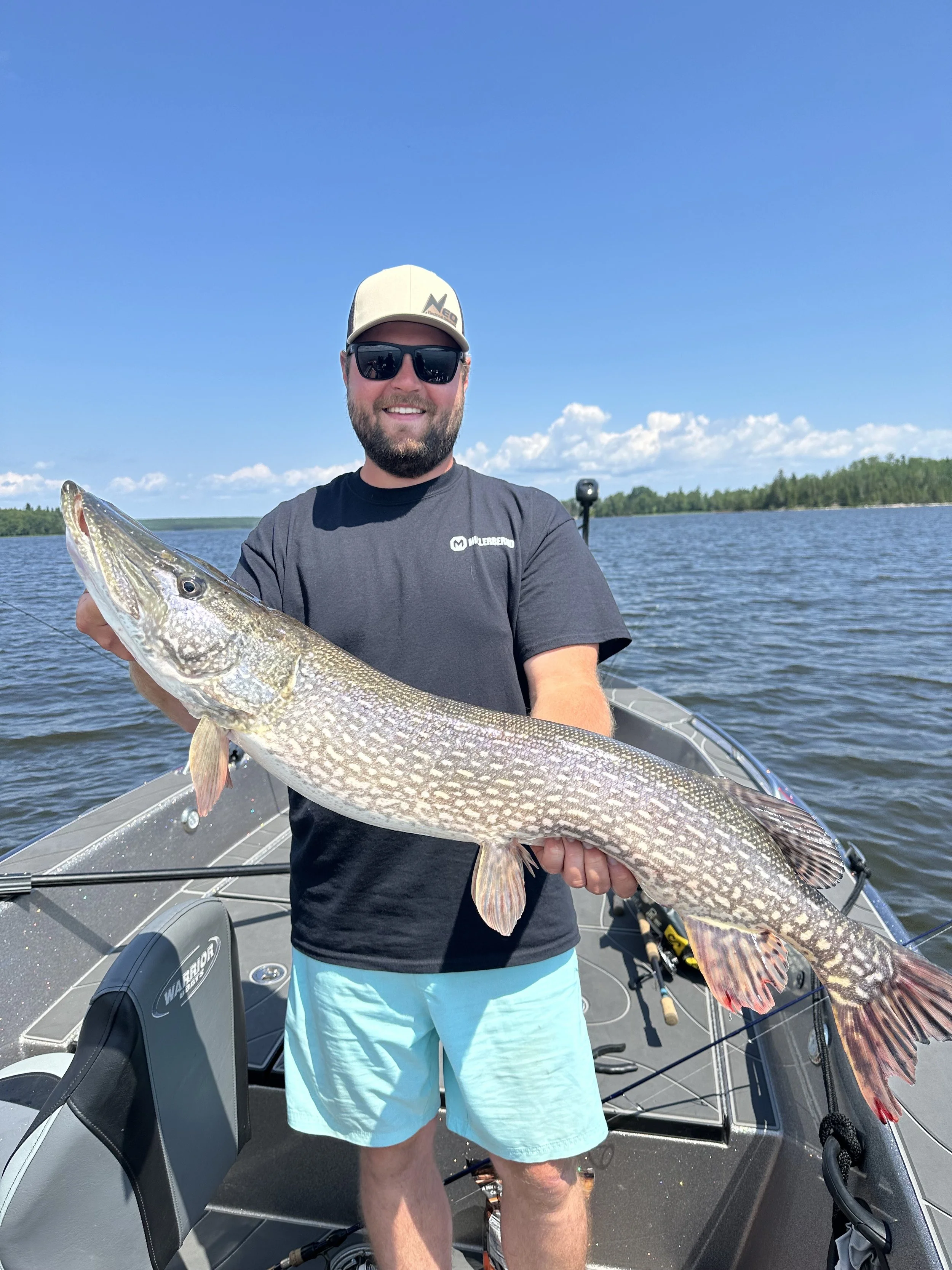 A man wearing a black t-shirt, light blue shorts, a beige cap, and sunglasses, holding a large fish on a boat in the middle of a lake under a blue sky with some clouds.