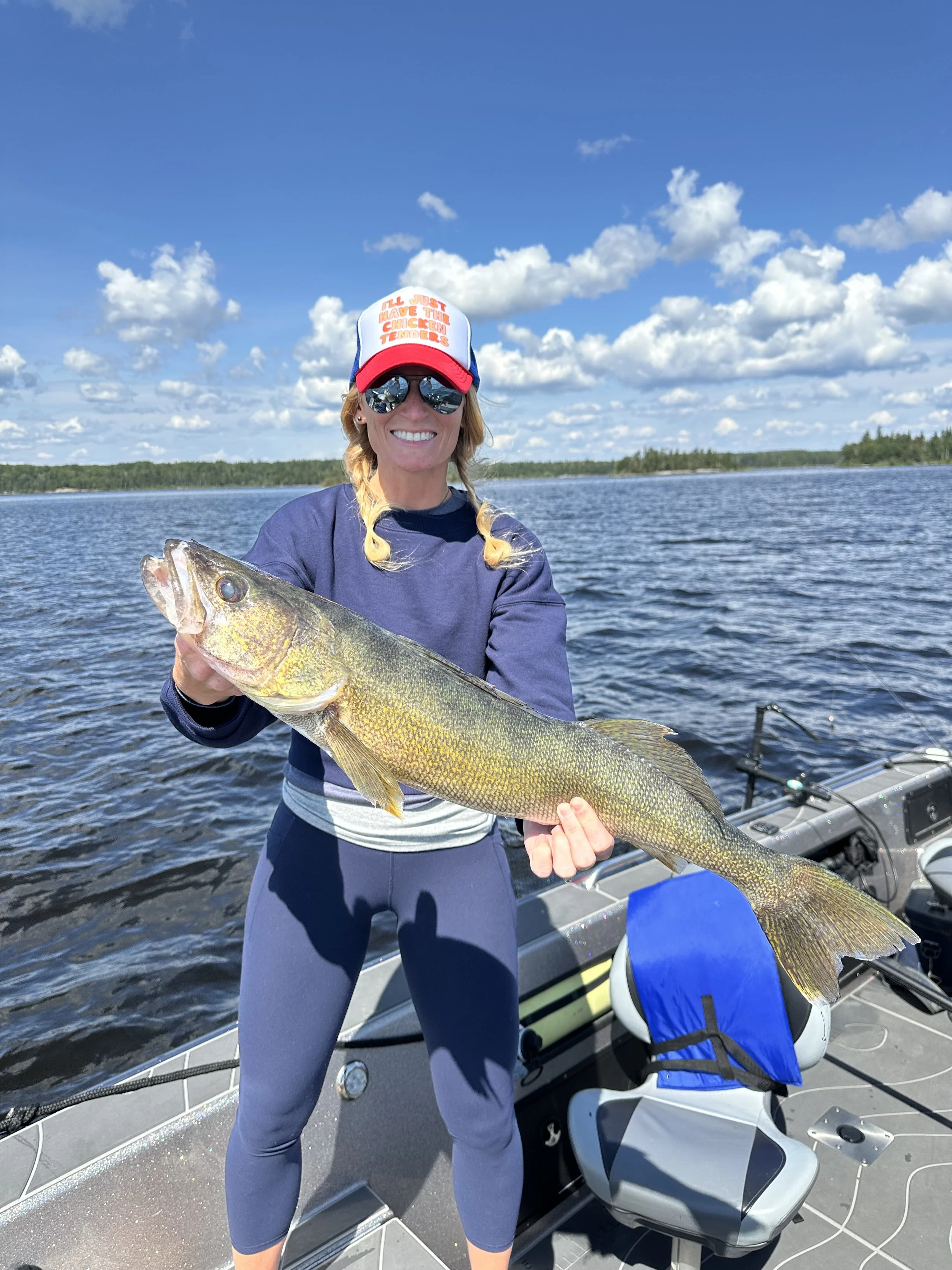Woman on a boat holding a large fish with a lake and blue sky with clouds in the background.