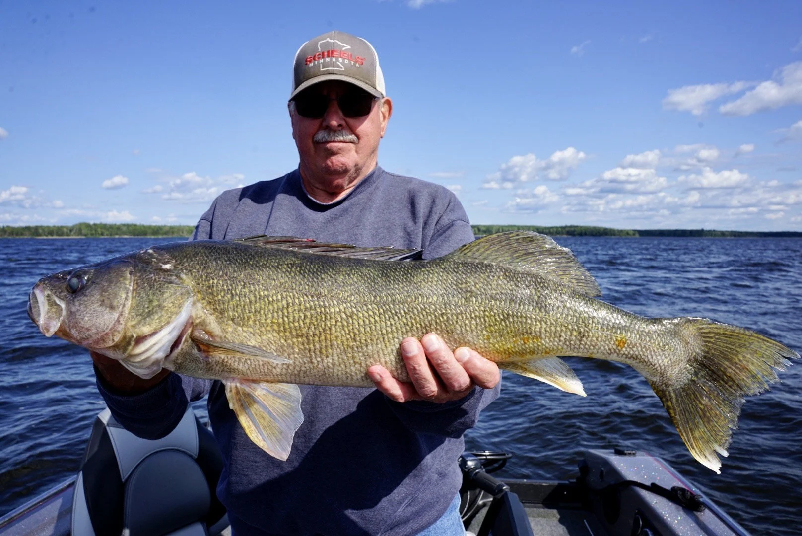 A man in a baseball cap, sunglasses, and a sweatshirt holding a large fish on a boat on a lake with a blue sky and clouds in the background.