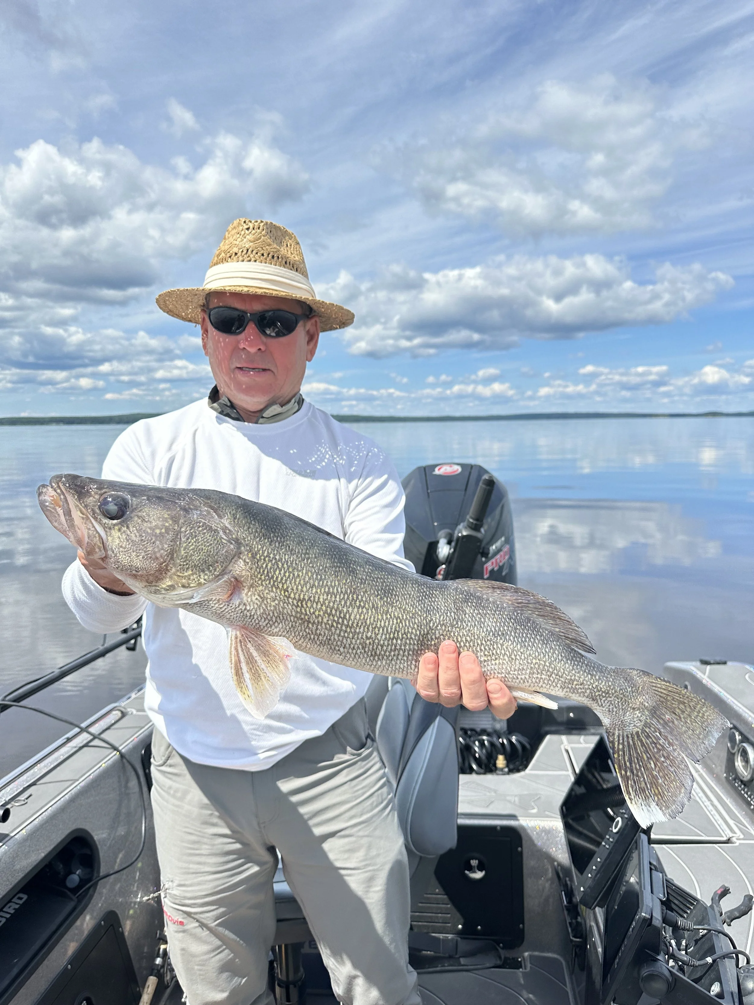 Man wearing sunglasses, a wide-brim hat, and a white shirt holding a large fish on a boat in a calm lake with a blue sky and clouds.