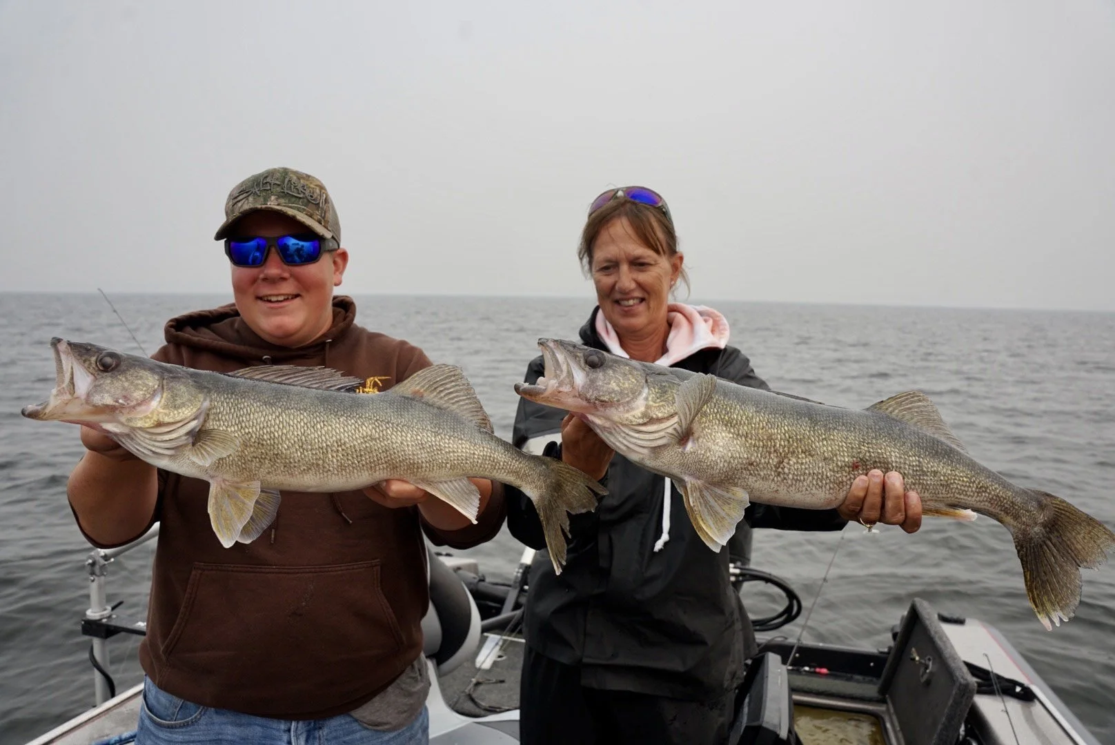 Two people holding large fish on a boat in open water, smiling, with an overcast sky.