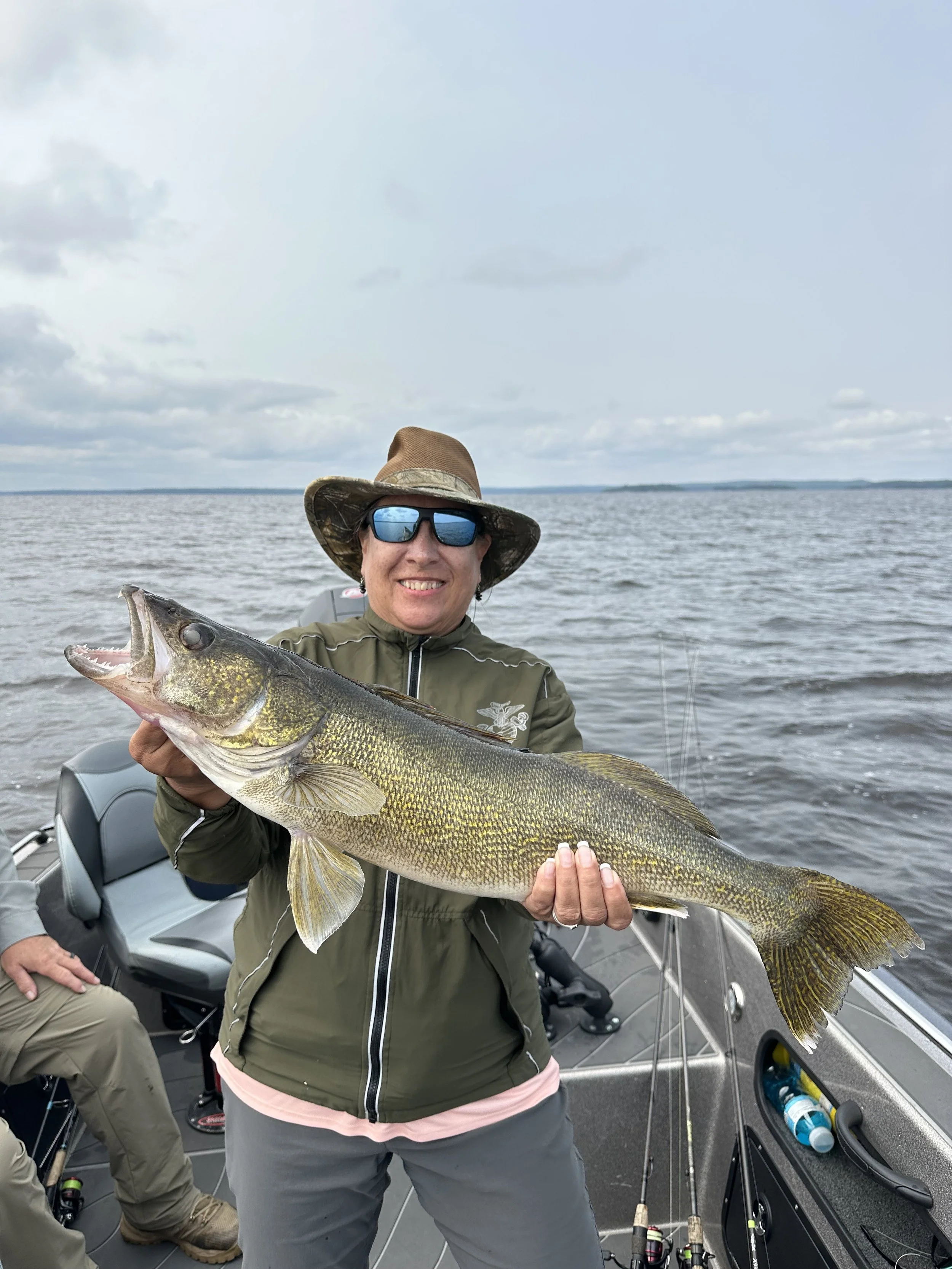 Woman wearing sunglasses and a hat on a boat holding a large fish she caught, with water and cloudy sky in the background.