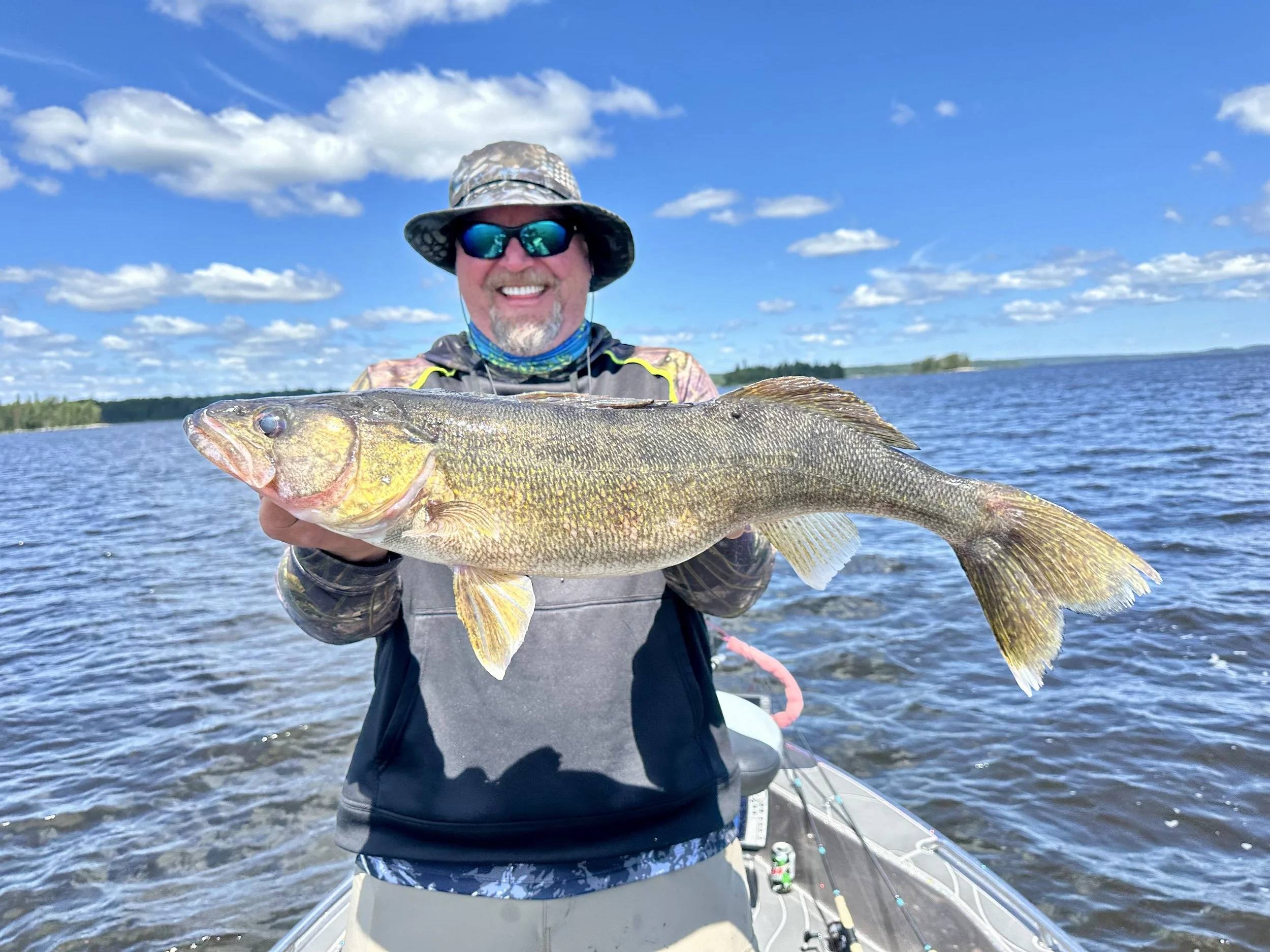 A man wearing sunglasses, a broad-brimmed hat, and fishing gear holding a large fish on a boat in a lake with a blue sky and scattered clouds.