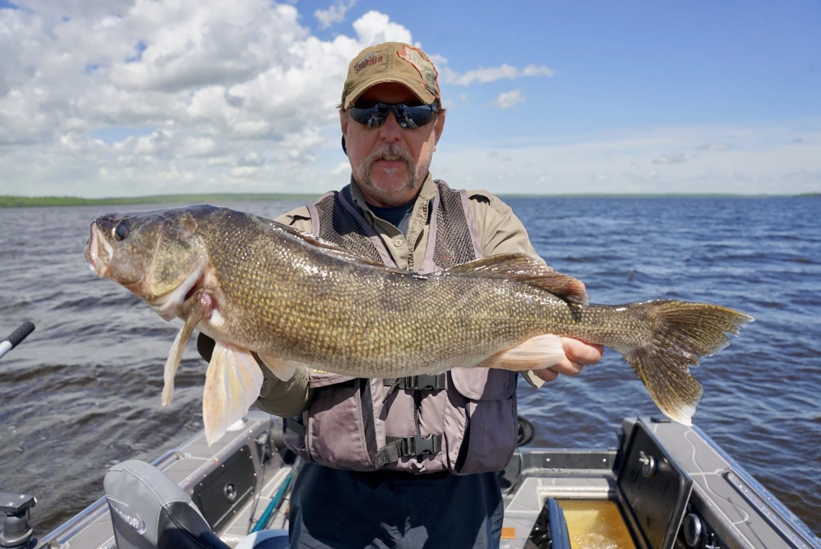 Man wearing sunglasses and a cap holding a large fish on a boat in a lake with cloudy sky.