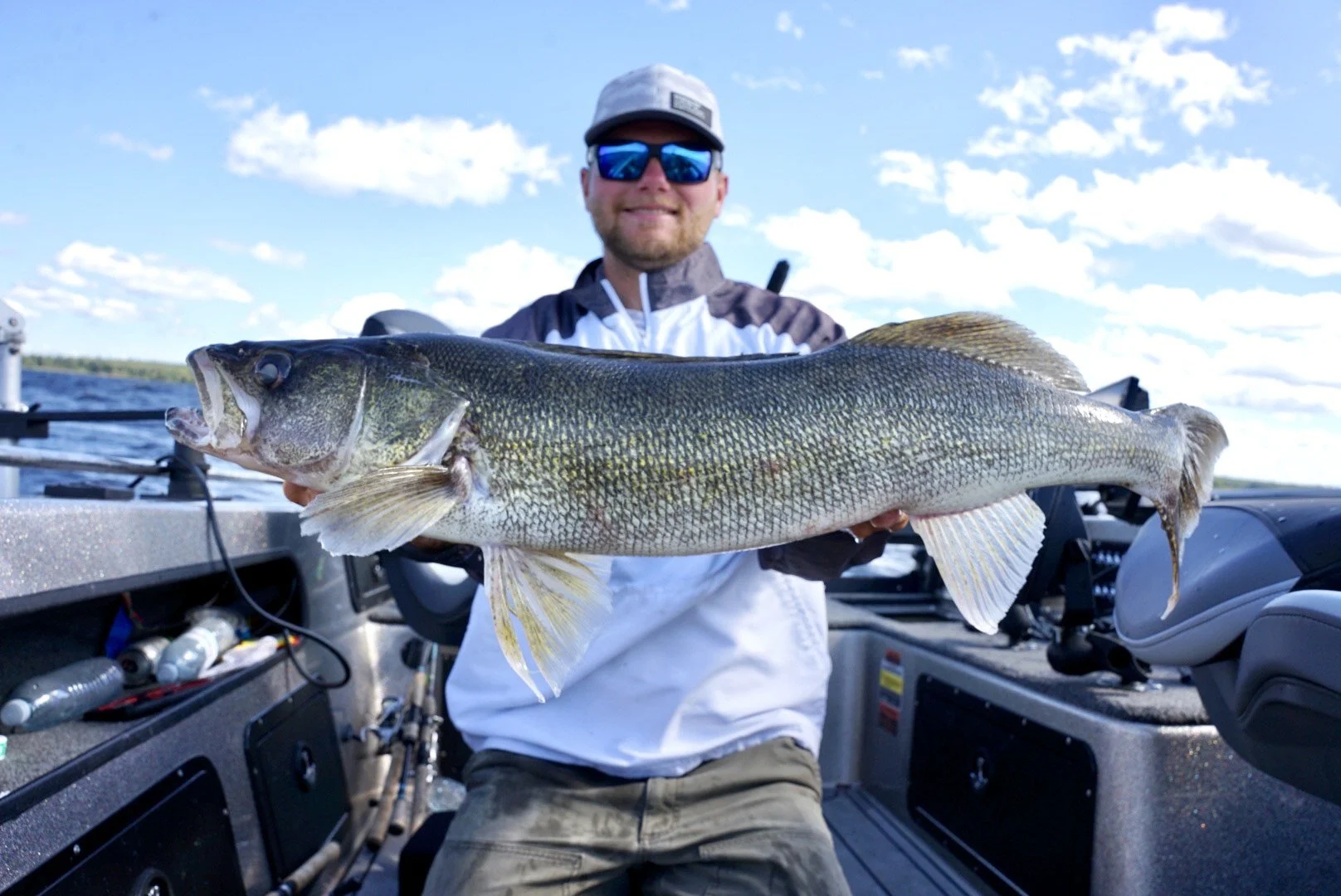 Man on a boat holding a large fish with a lake and blue sky in the background.