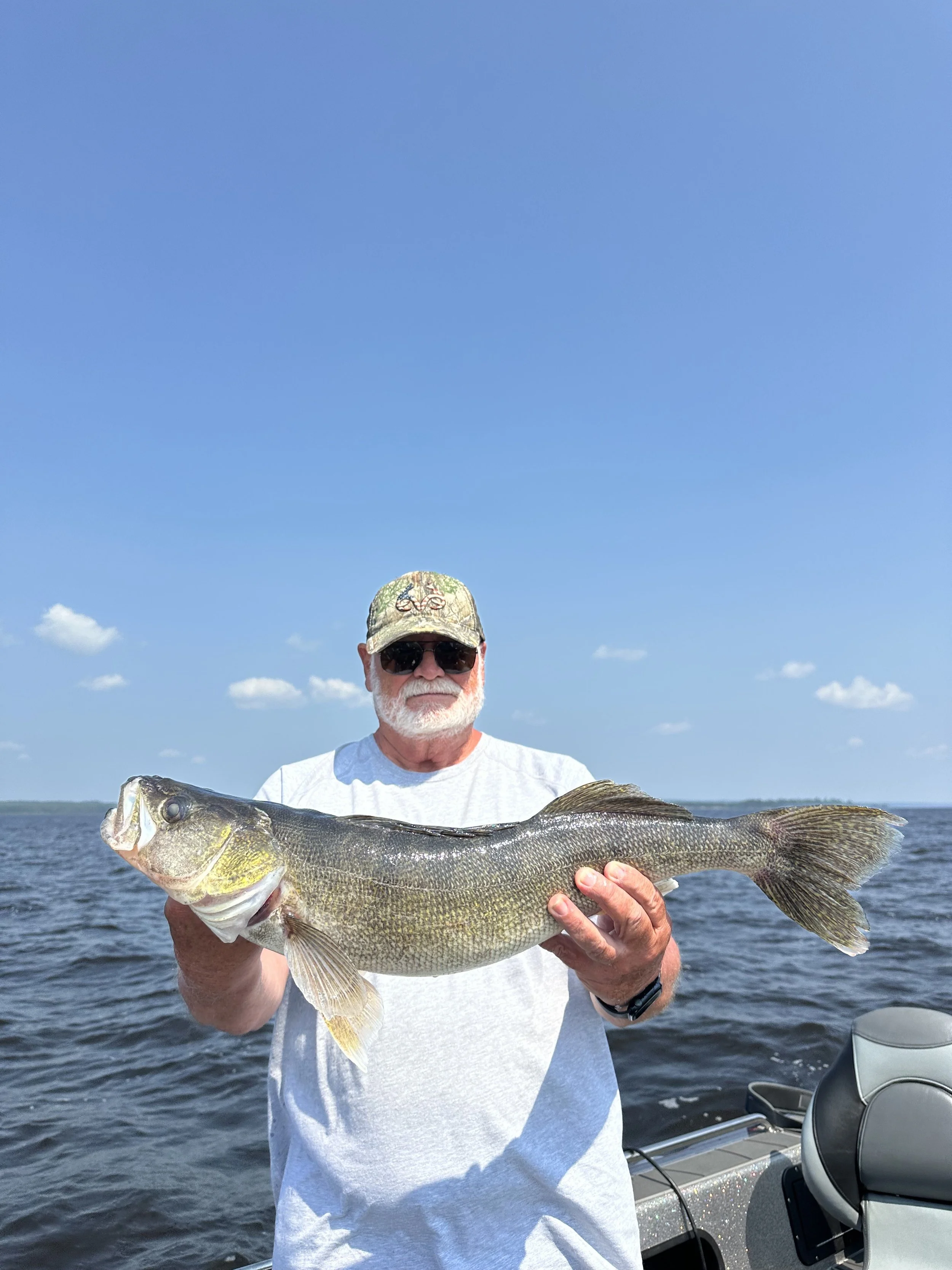 An older man with a white beard and sunglasses holding a large fish on a boat in the water on a sunny day.