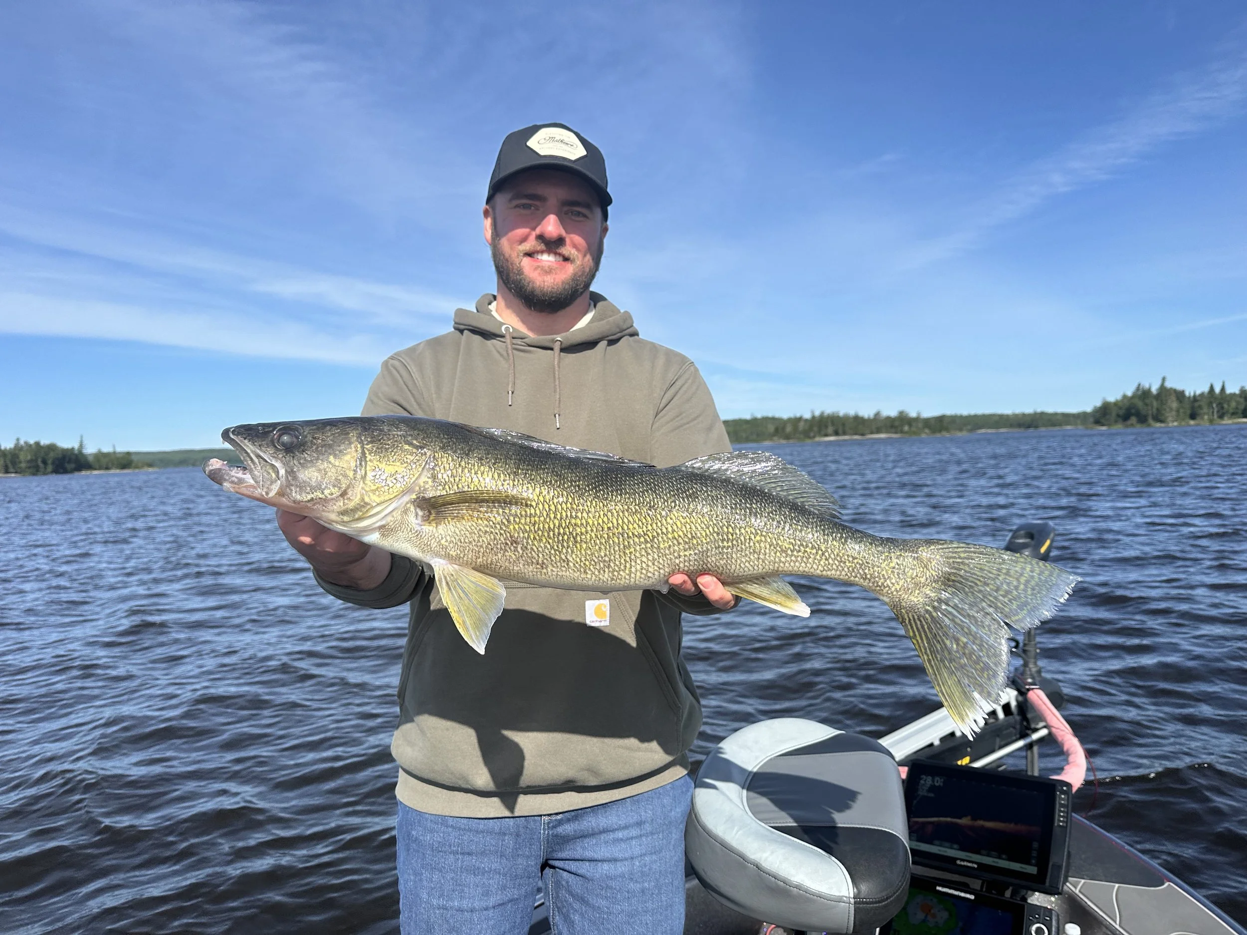A man smiling and holding a large fish on a boat in a lake with trees in the background, under a clear blue sky.