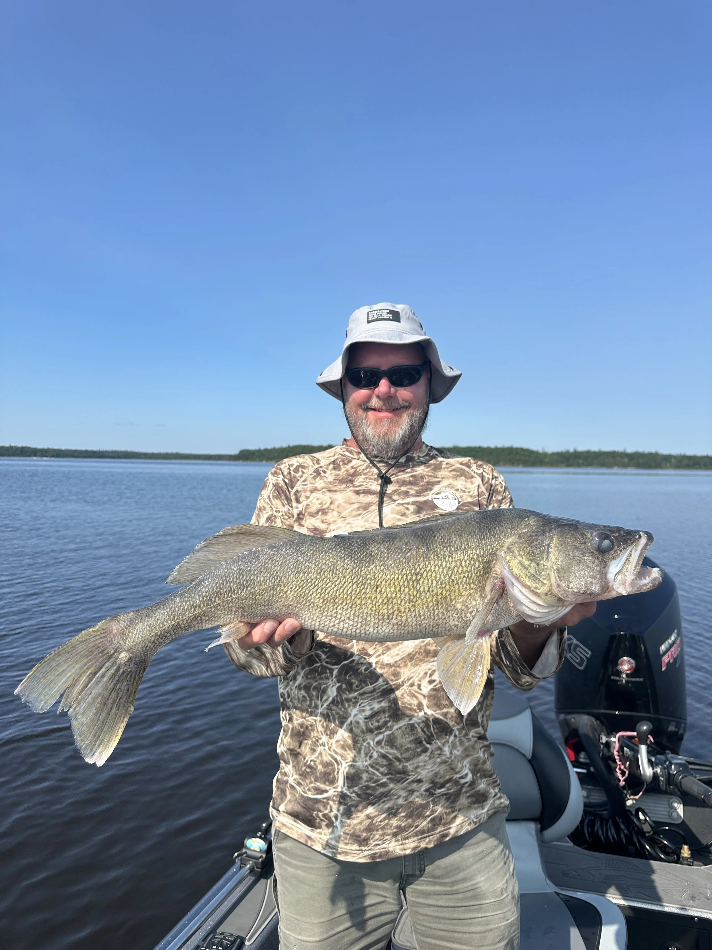 Man in camouflage shirt, sunglasses, and bucket hat holding a large fish on a boat with a calm body of water and a clear blue sky in the background.