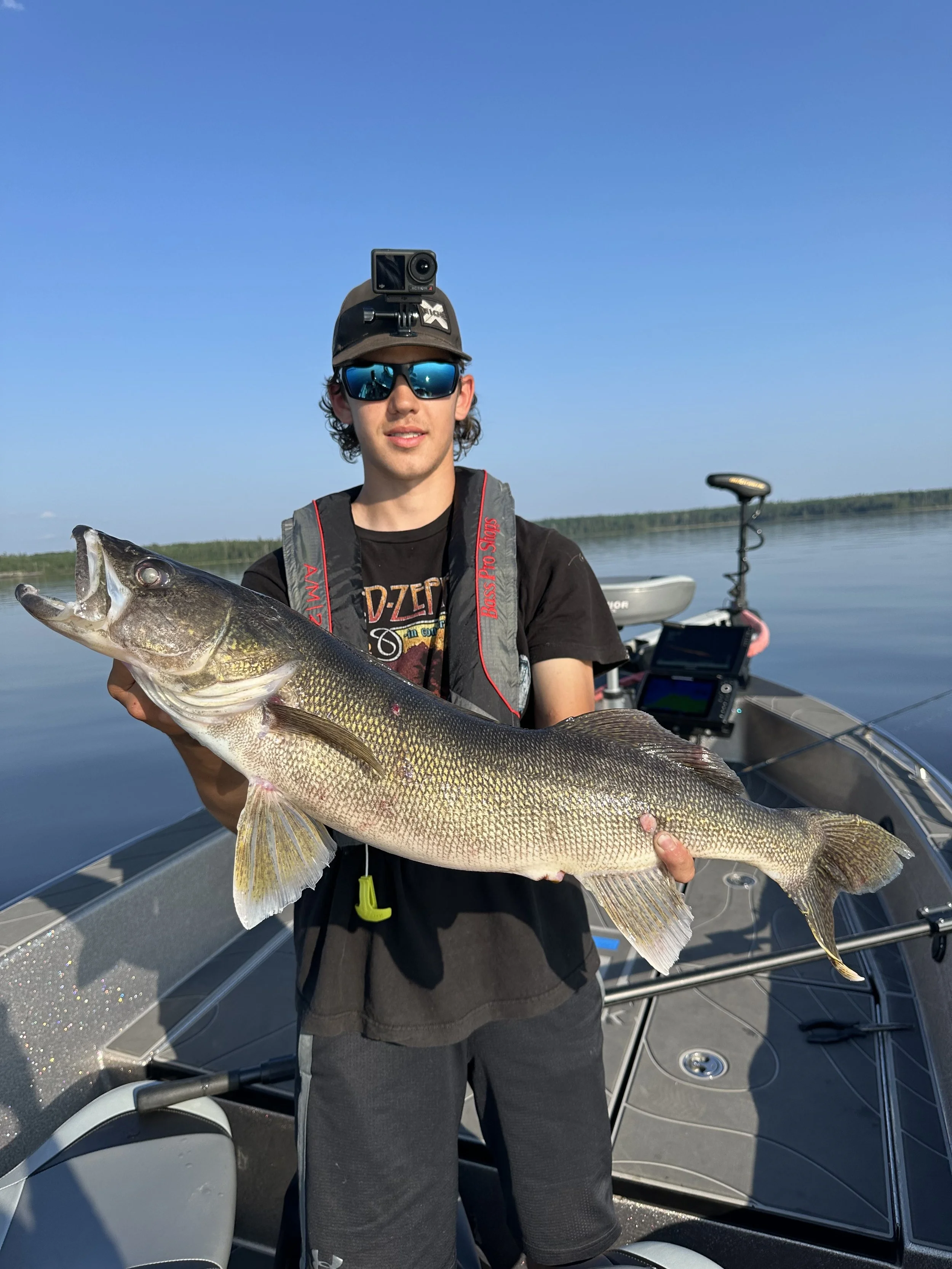 Young man holding a large fish on a boat in a calm lake, wearing sunglasses, a cap with a camera, and a life jacket.