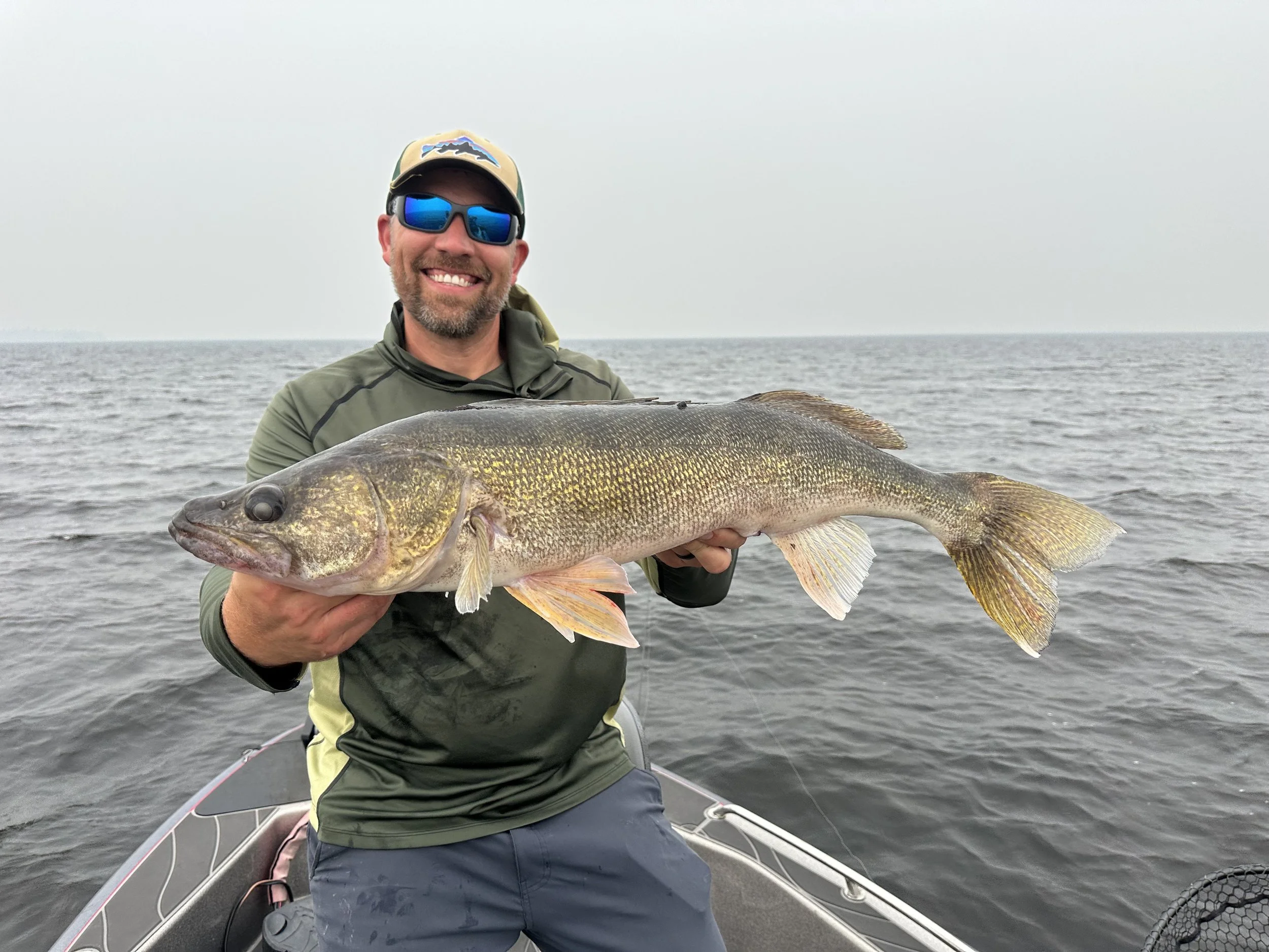 A man wearing sunglasses and a cap smiling while holding a large fish on a boat in open water.