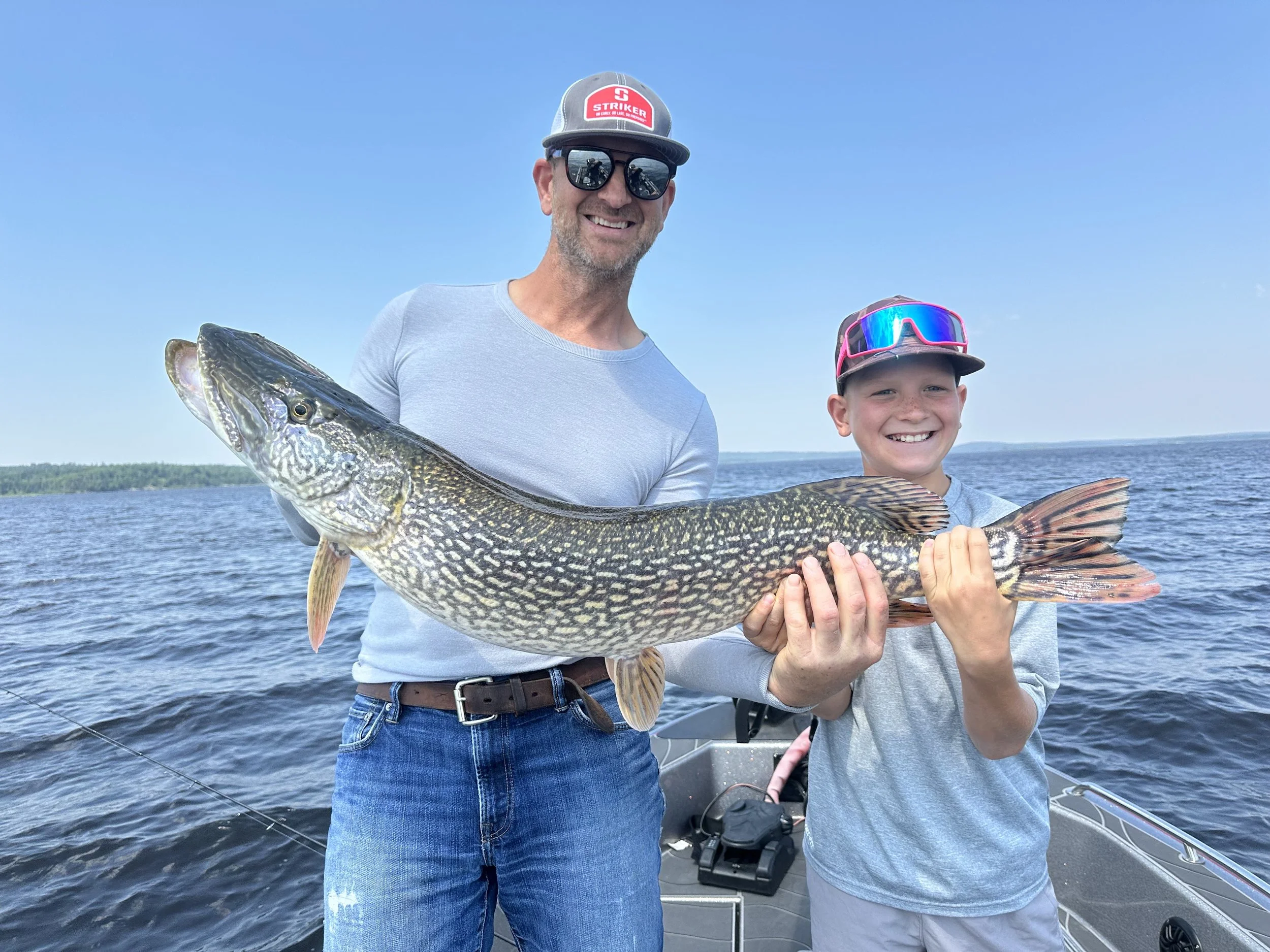 A man and a boy on a boat in the water holding a large fish, with the man smiling and wearing sunglasses and a baseball cap, and the boy smiling and wearing sunglasses and a cap, with a scenic water view in the background.