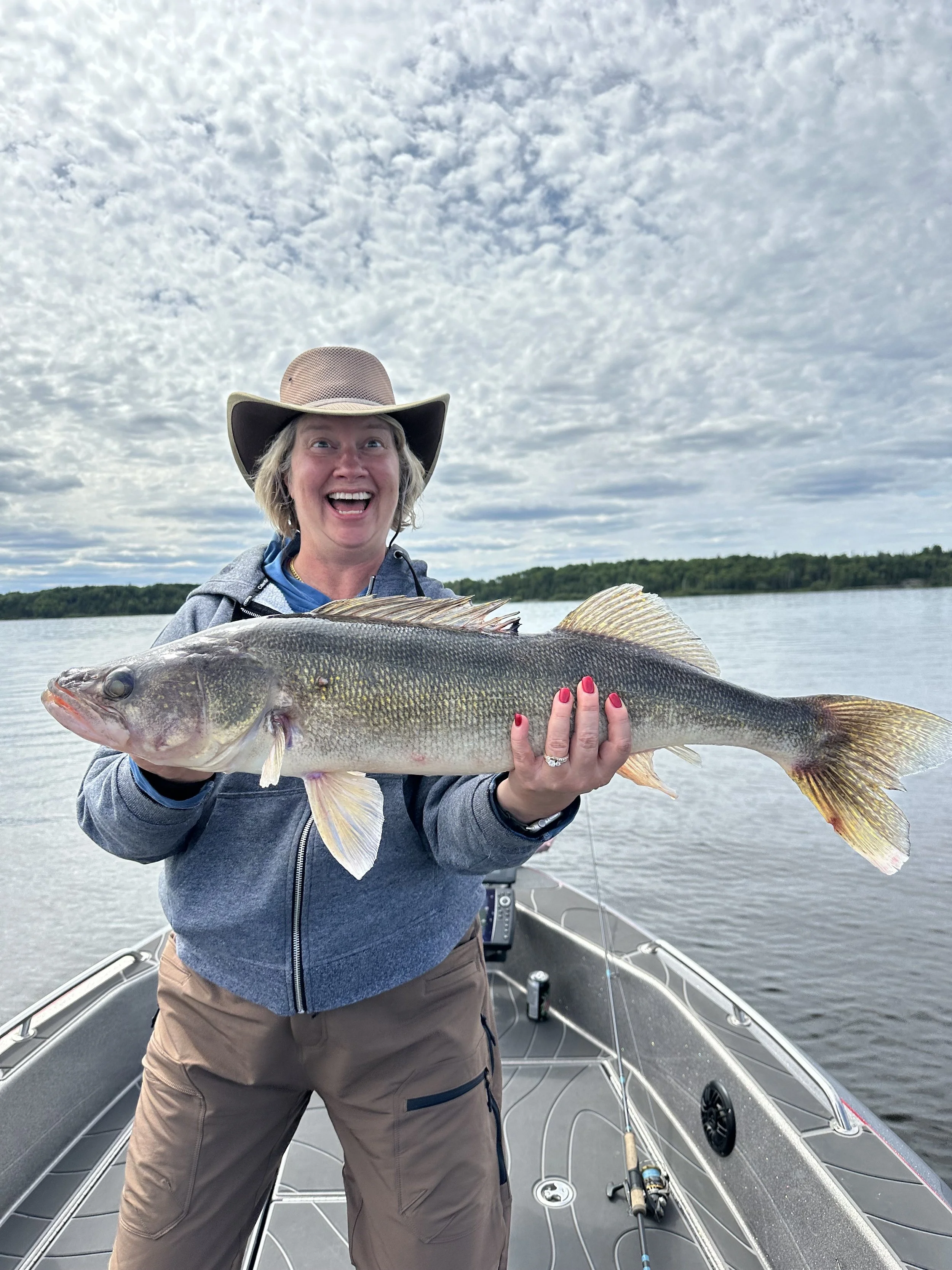 Woman holding a large fish on a boat on a lake during daytime, smiling and wearing a wide-brimmed hat and casual outdoor clothing.