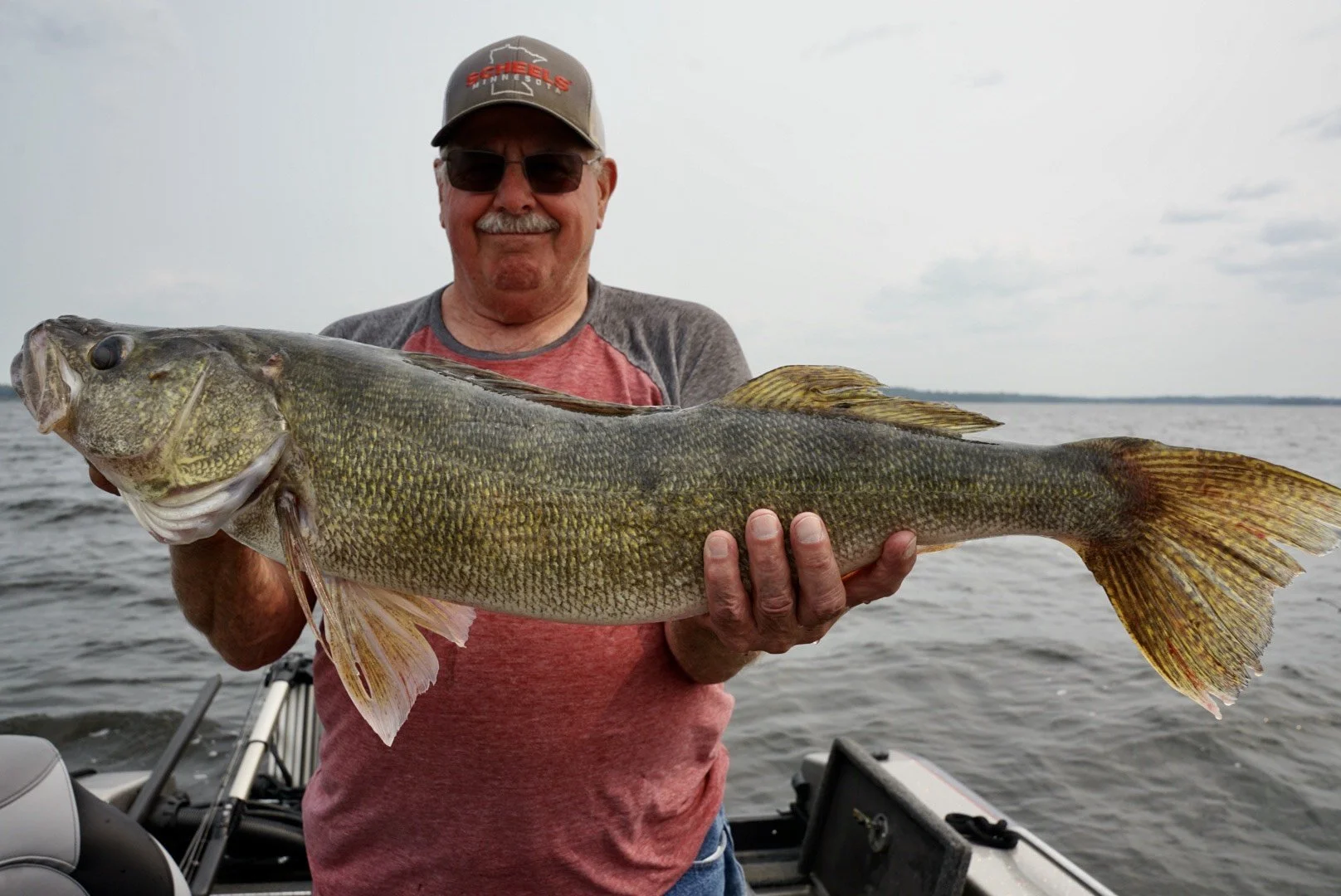 A man wearing sunglasses and a cap holding a large fish on a boat in a body of water.