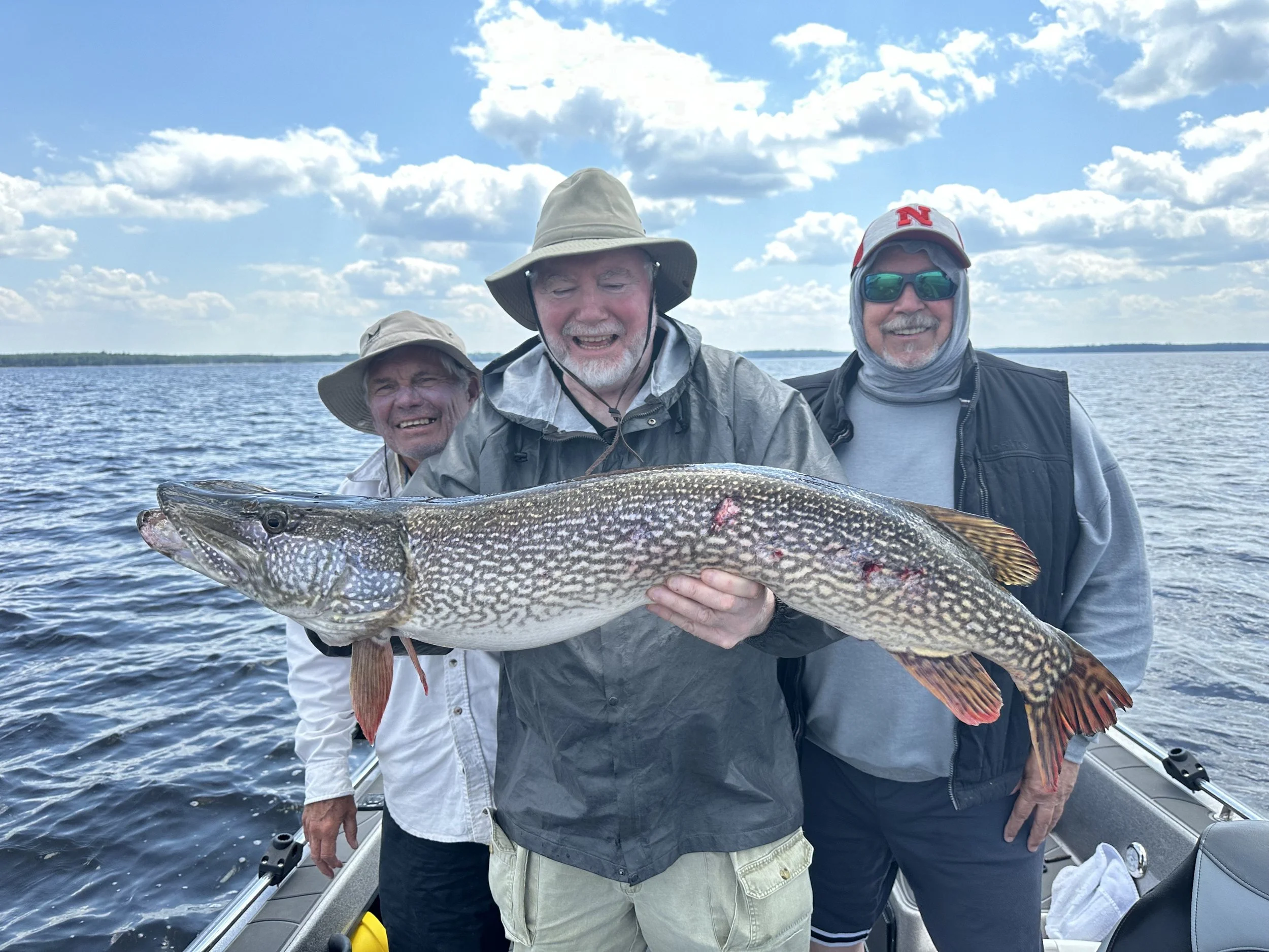 Three men on a boat holding a large fish, with water and a partly cloudy sky in the background.