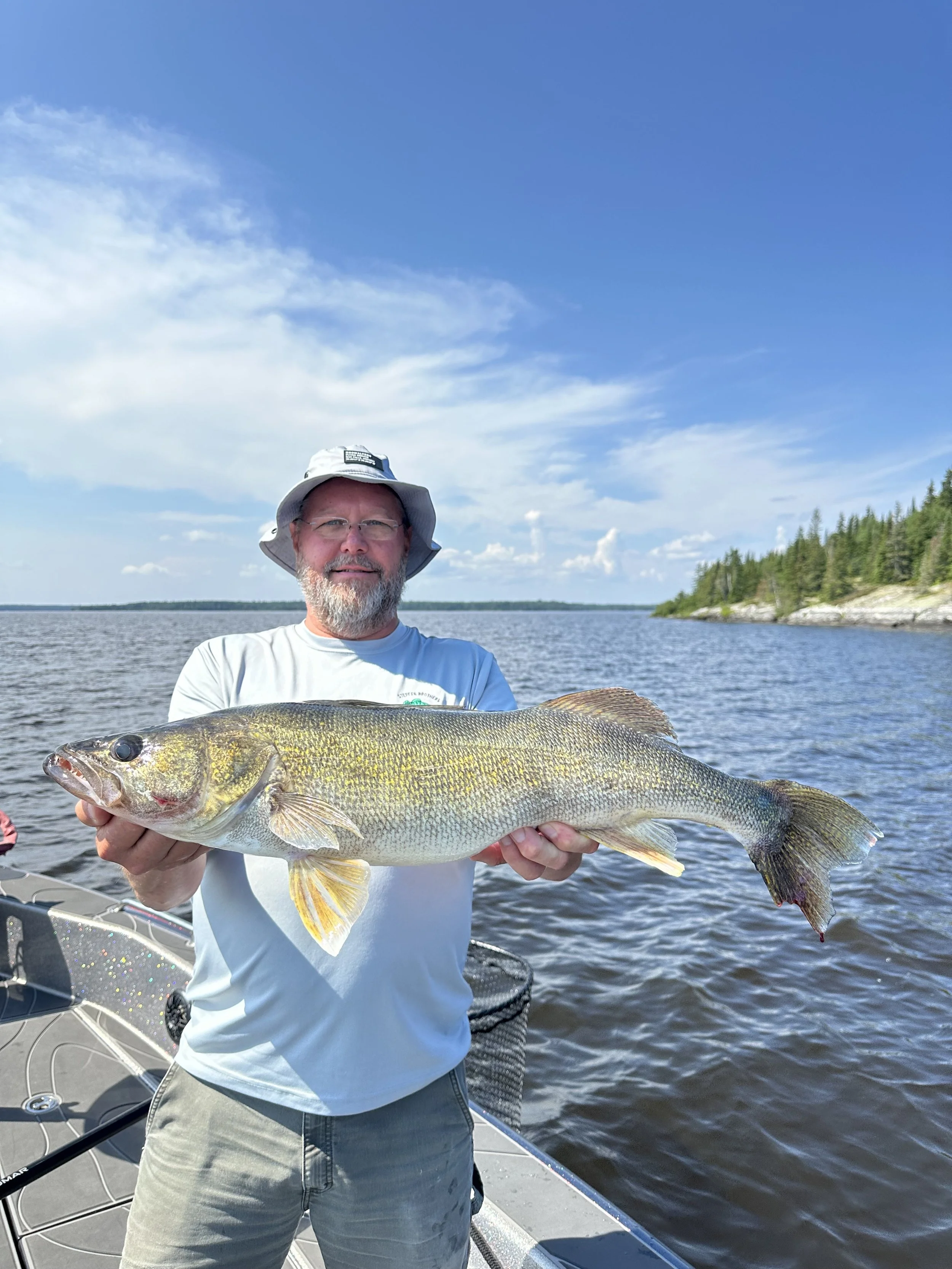A man wearing a white shirt, gray shorts, and a gray hat holding a large fish on a boat in a lake.