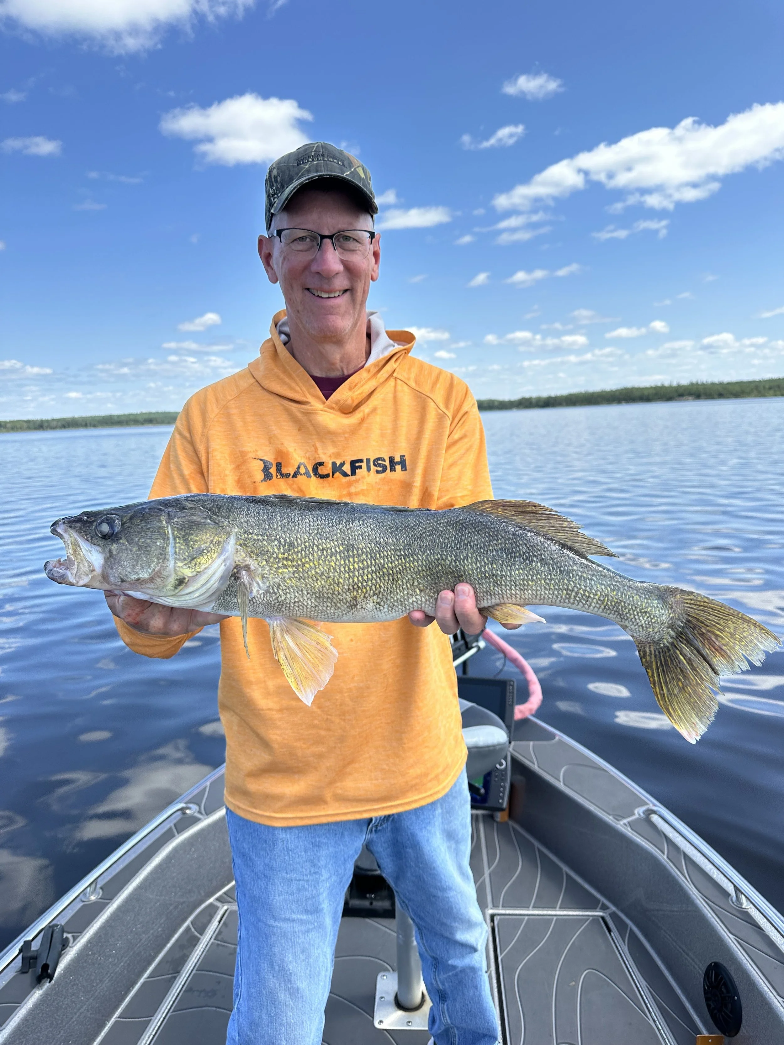 A man smiling on a boat holding a large fish against a backdrop of a calm lake and blue sky with scattered clouds.