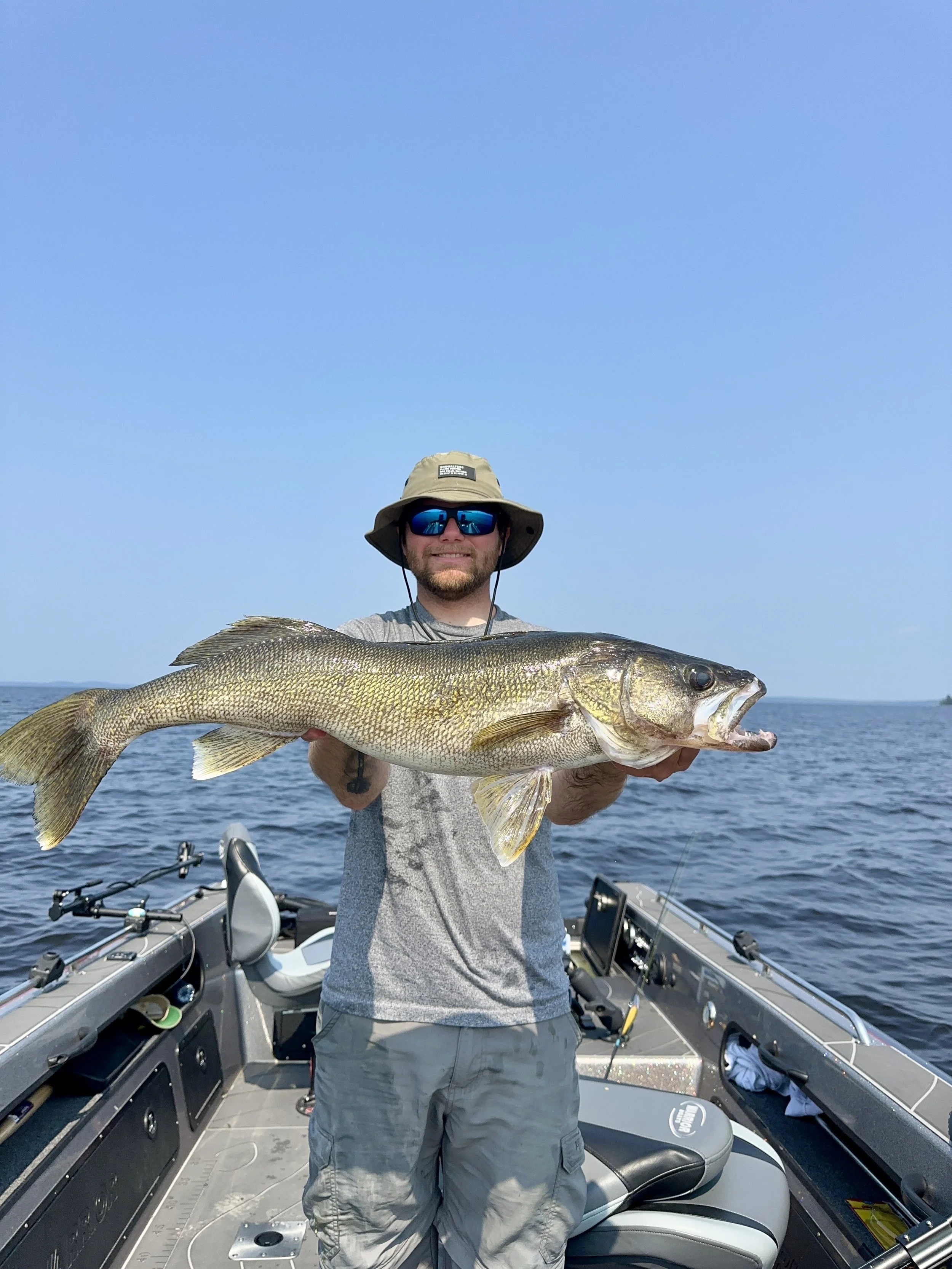 Man in gray t-shirt, khaki shorts, and wide-brim hat holding a large fish on a boat in open water with clear blue sky.