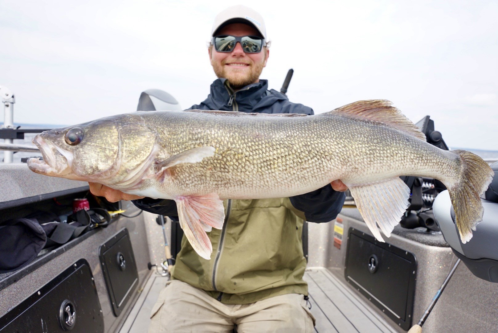 A man wearing sunglasses, a cap, and a jacket on a boat holding a large fish with both hands.