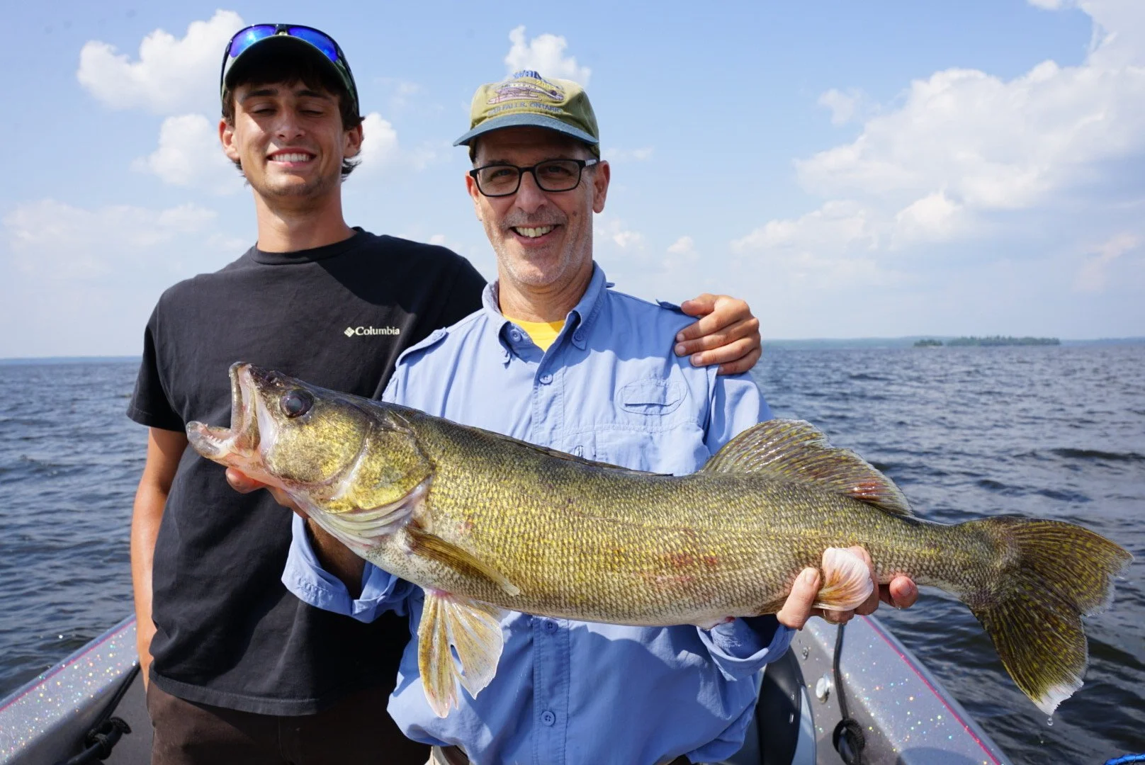 Two men on a boat in the water, one holding a large fish they caught, both smiling, with a partly cloudy sky in the background.