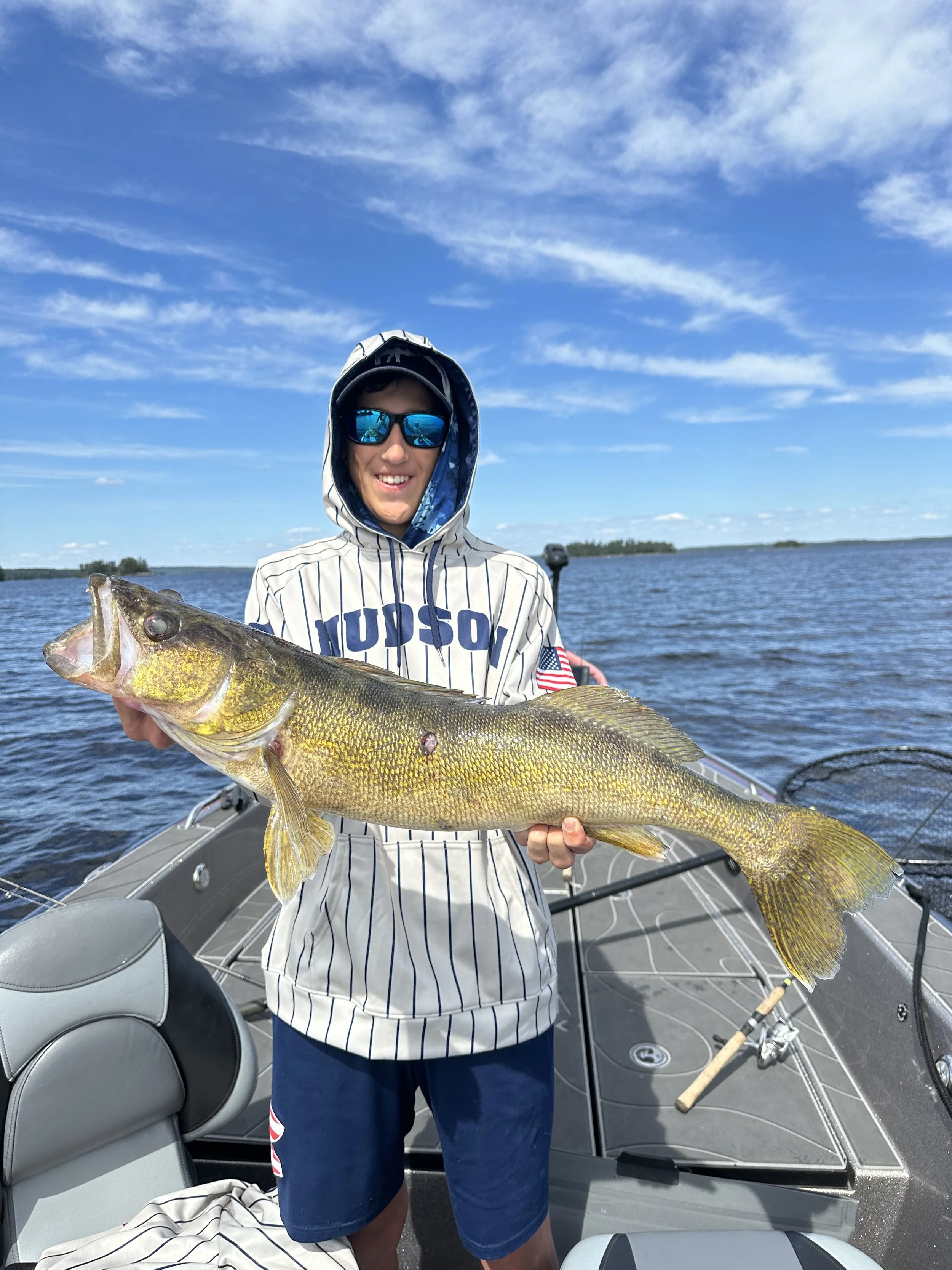 A young man wearing sunglasses, a hoodie, and shorts, smiling while holding a large fish on a boat in a lake under a blue sky.