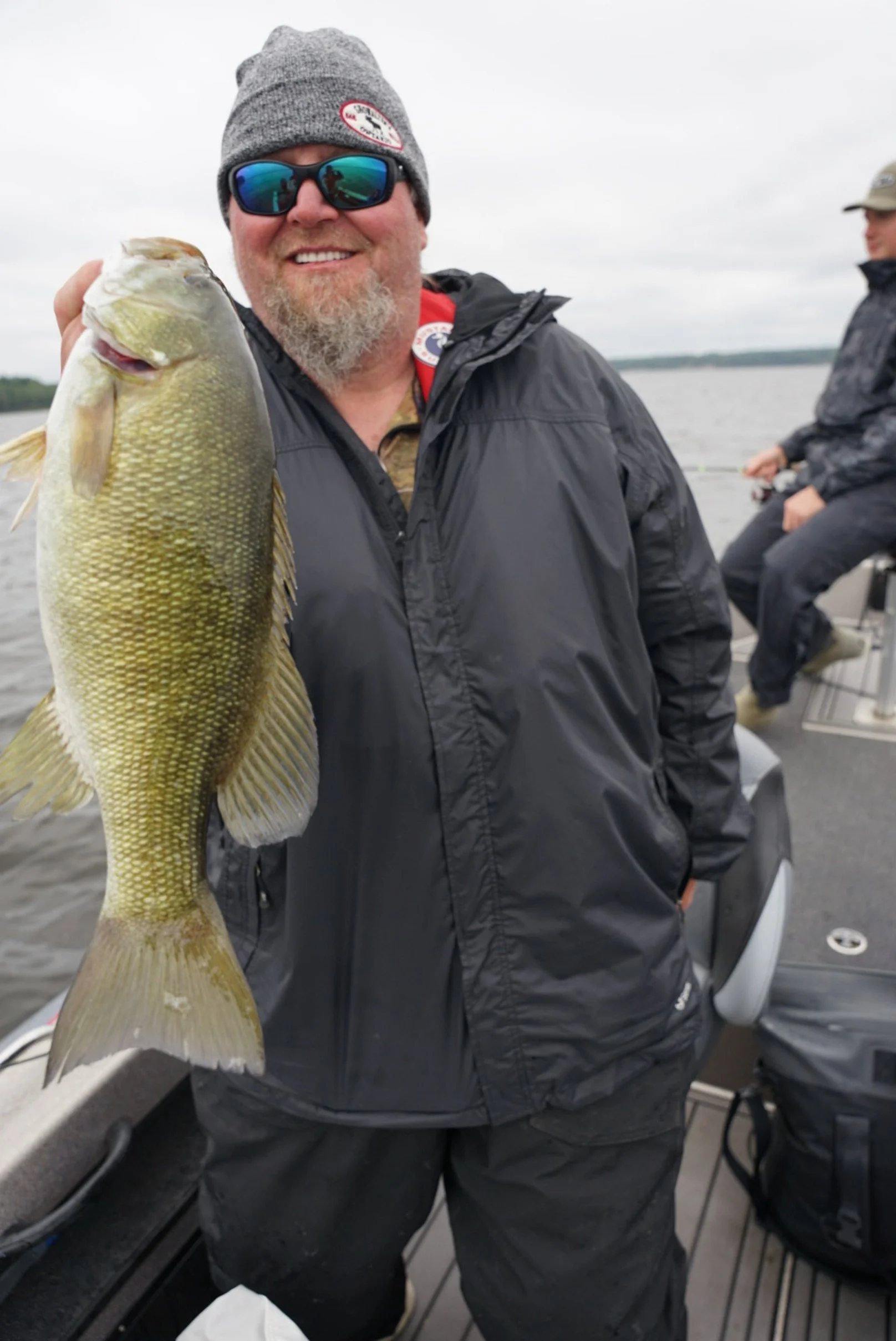 Man holding a large fish on a boat during cloudy weather, with another person fishing in the background.