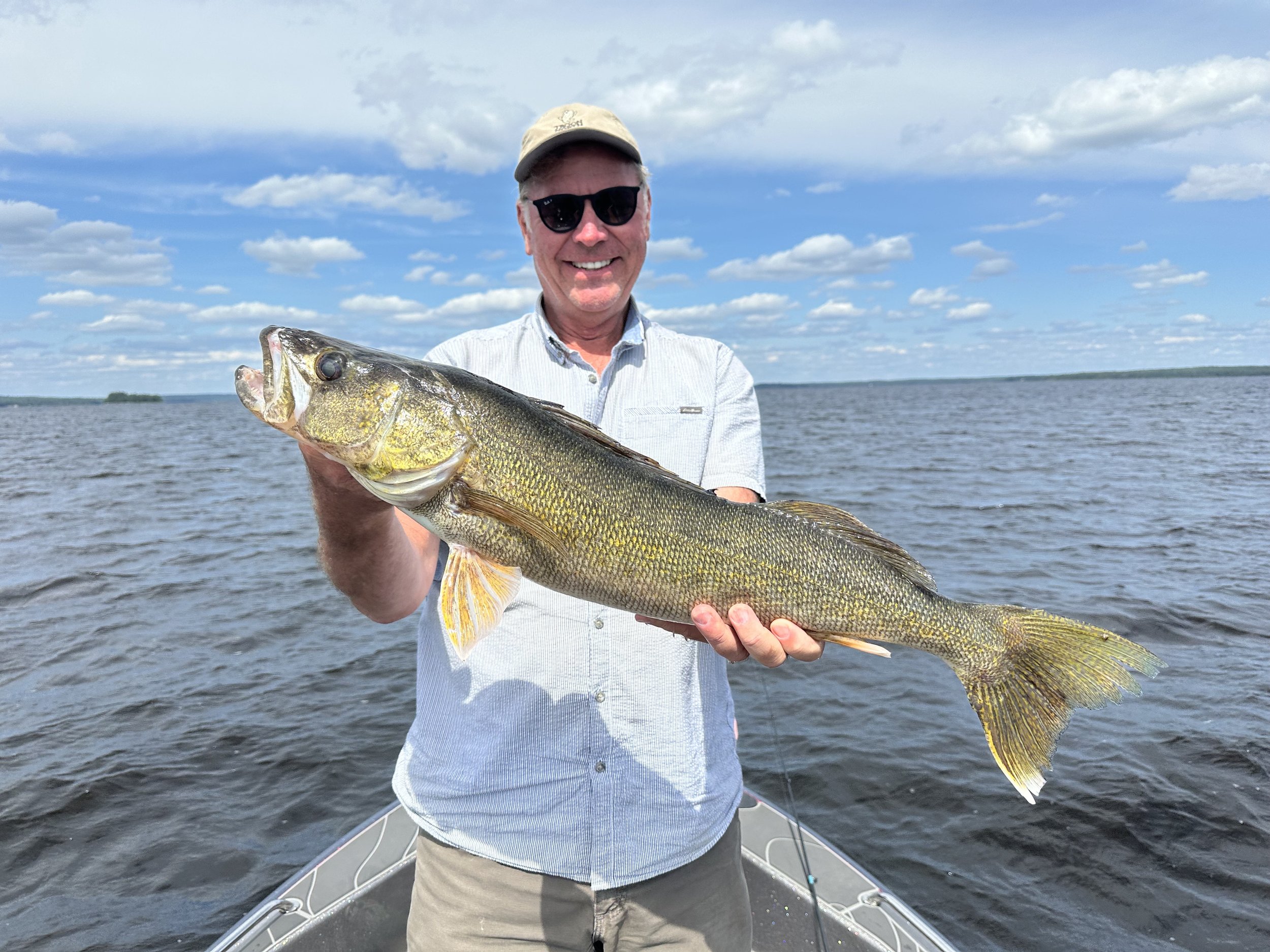 Man wearing sunglasses and a light-colored hat holding a large fish on a boat in a lake with a partly cloudy sky.