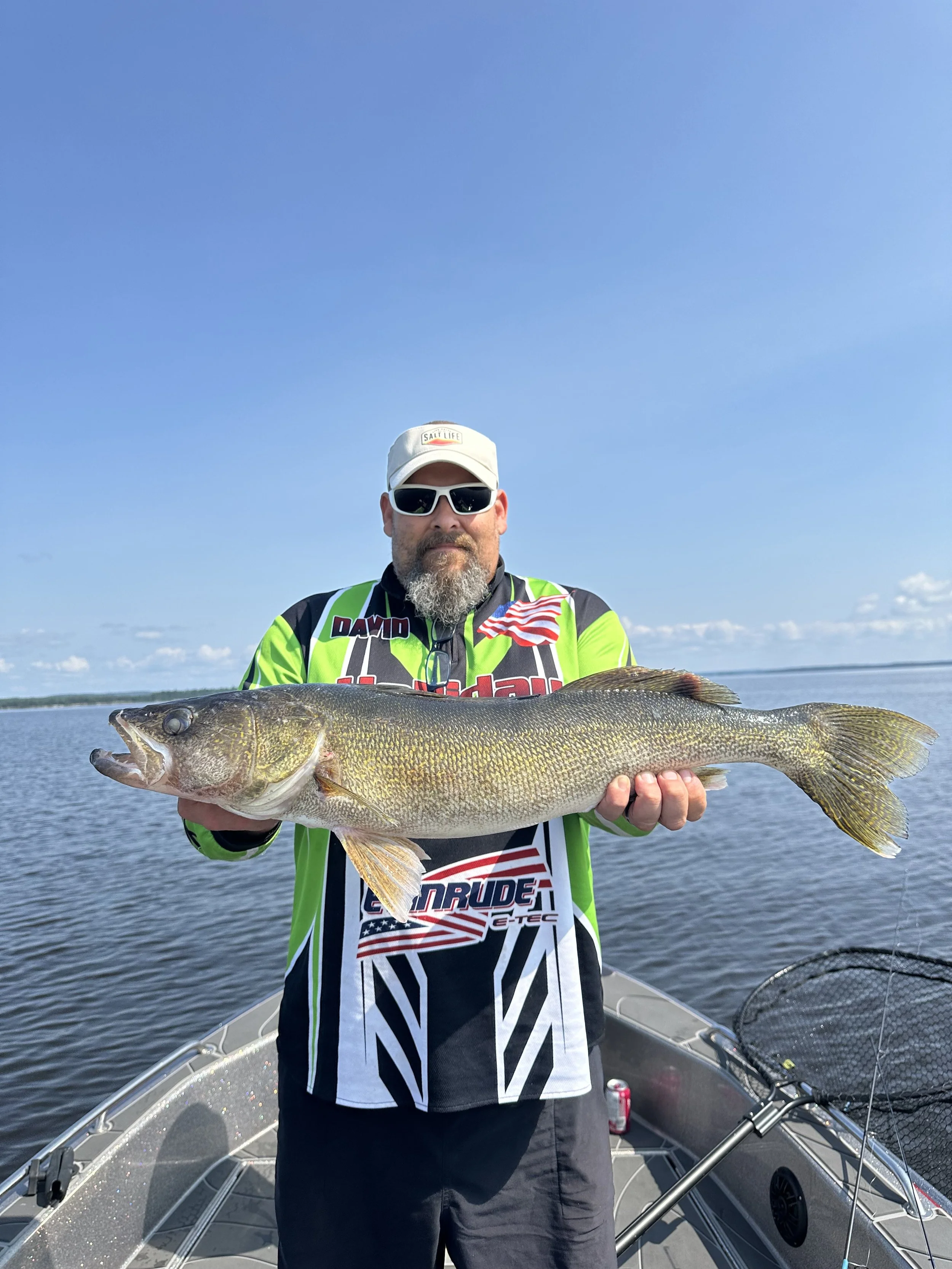 A man wearing sunglasses, a white cap, and a green, black, and white fishing shirt stands on a boat holding a large bass fish with both hands against a lake and blue sky background.