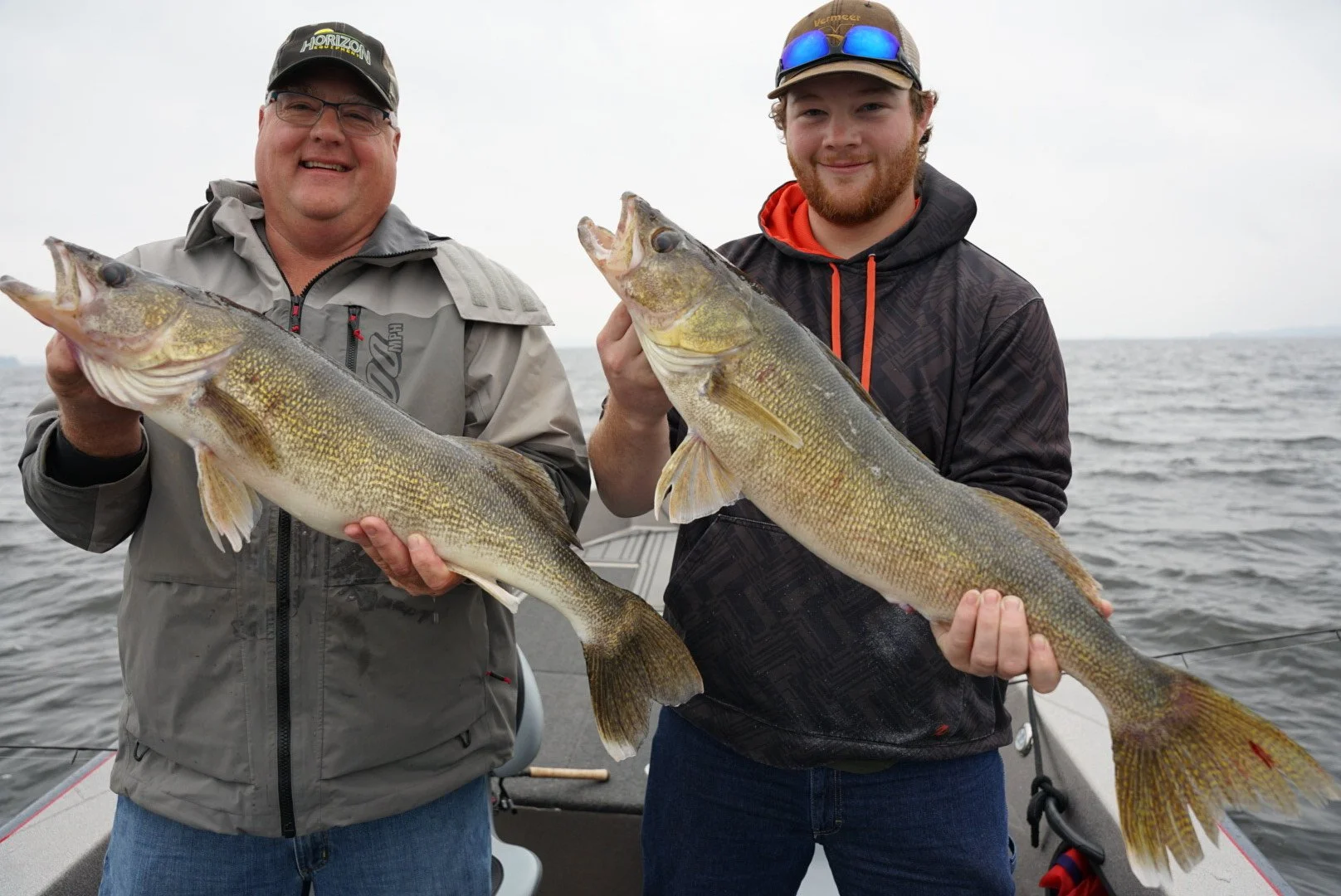 Two men on a boat holding large fish they caught, with water and cloudy sky in the background.