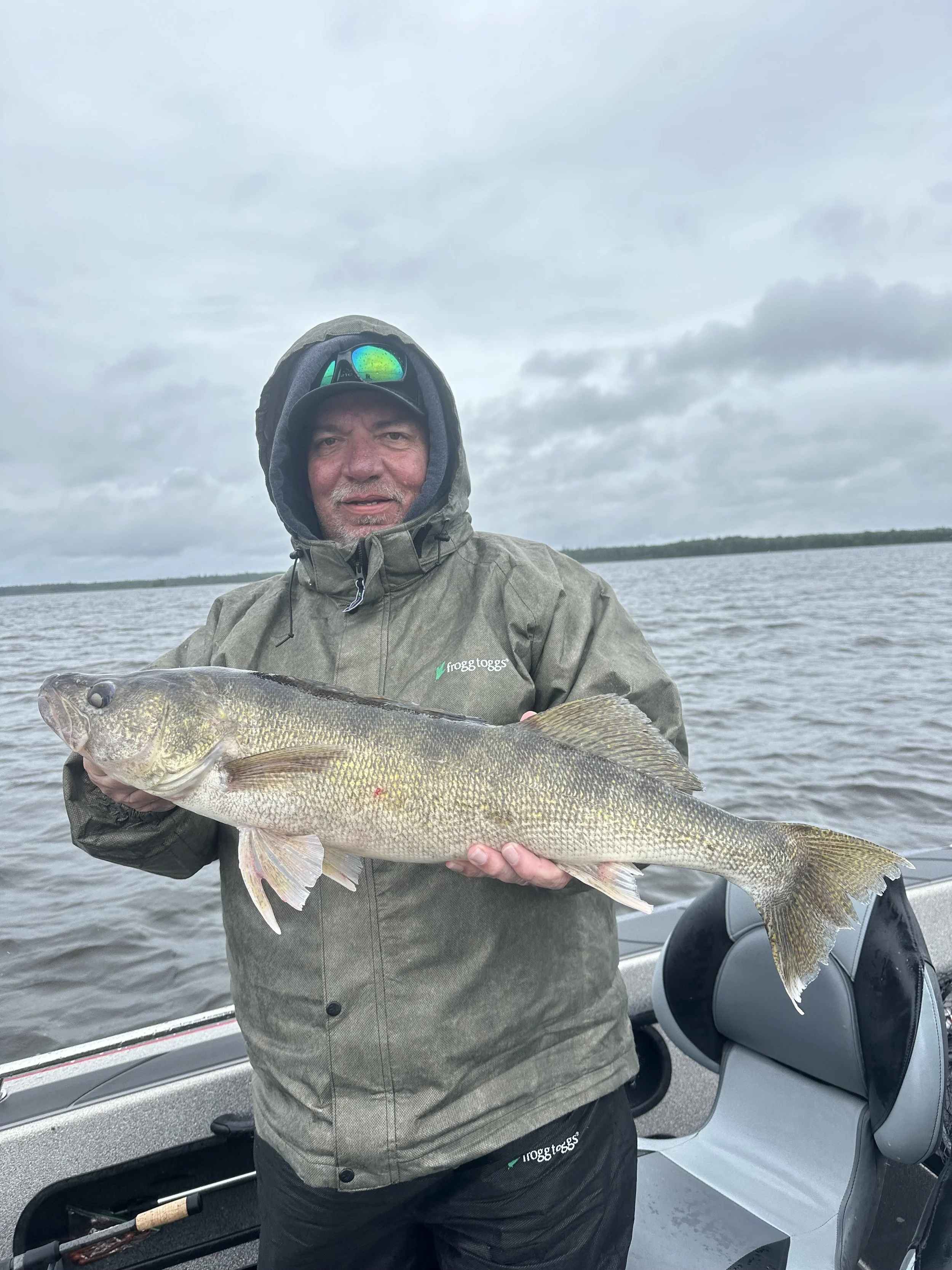 Man in rain jacket holding a large fish on a boat in a lake under cloudy sky.