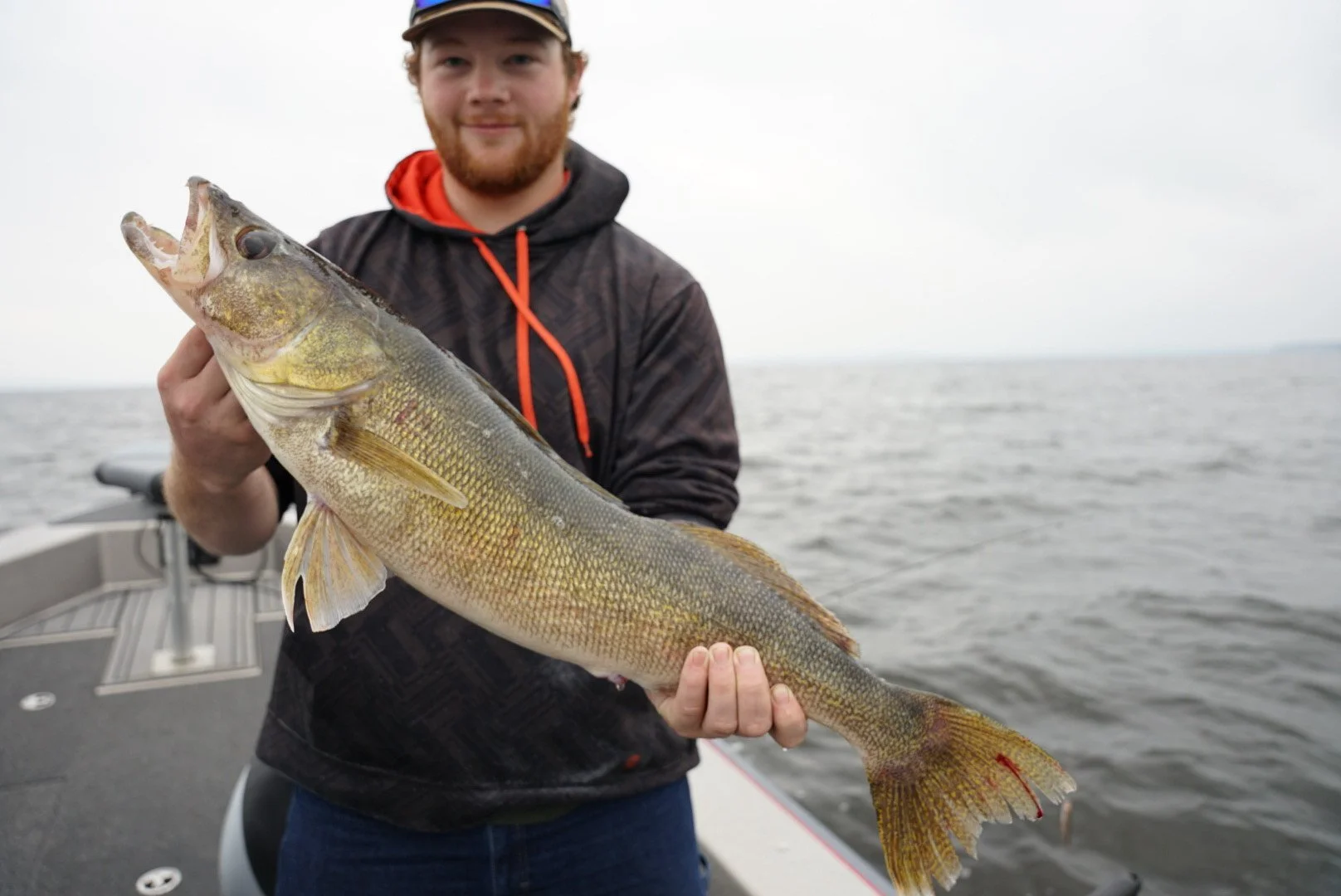 Man on a boat holding a large fish over a body of water with a cloudy sky, wearing a dark jacket and a cap.