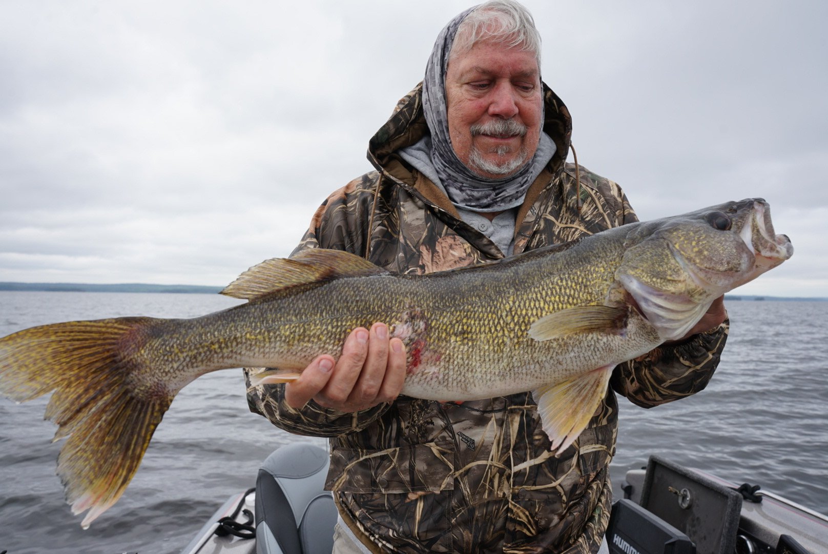 A man in camouflage clothing holding a large fish on a boat in a lake under a cloudy sky.