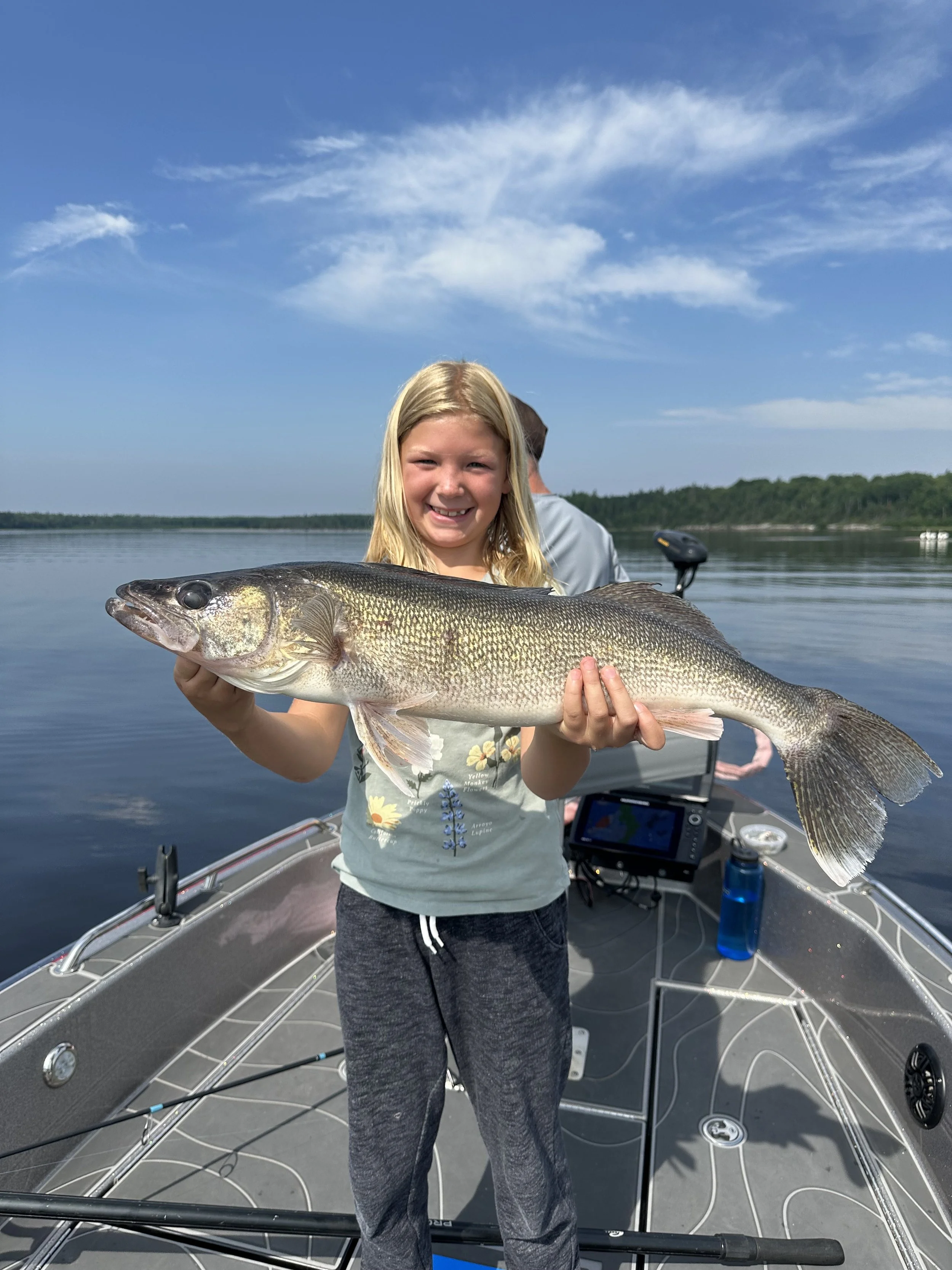 A young girl on a boat holding a large fish she caught, with water and trees in the background on a sunny day.