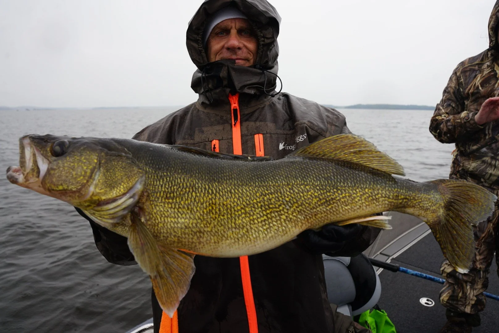 A man in waterproof clothing on a boat holding a large fish with yellow and greenish scales, dark fins, and an open mouth, over a body of water with a cloudy sky.