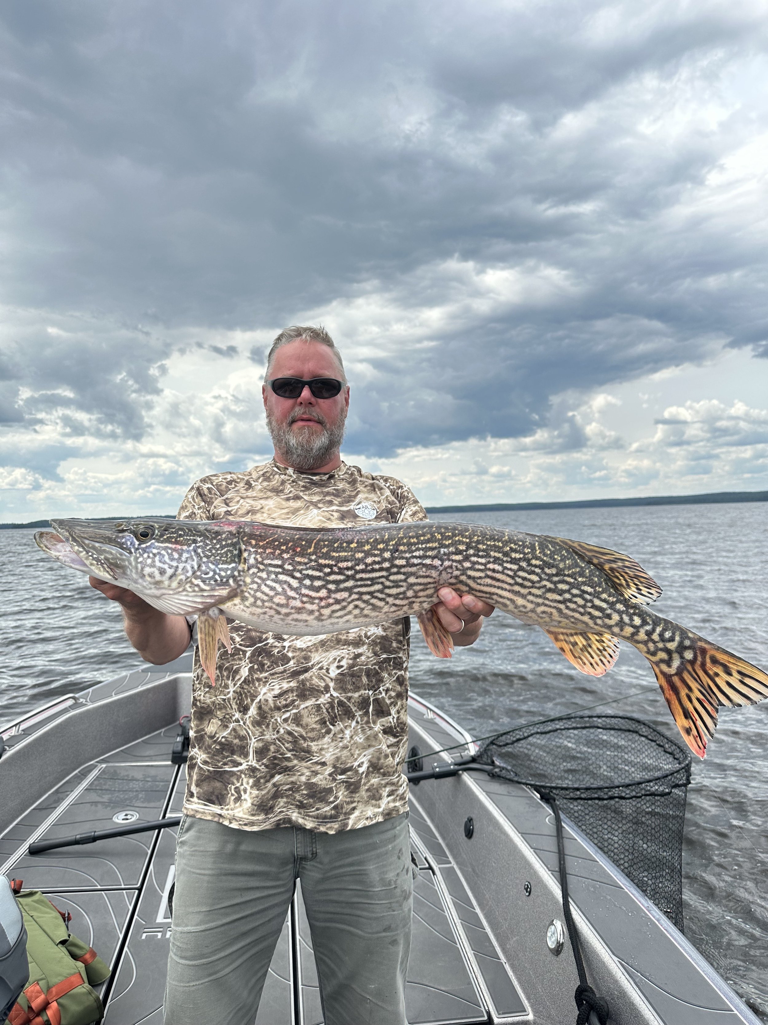 A man wearing sunglasses and camouflage shirt holding a large fish on a boat in a lake under cloudy sky.