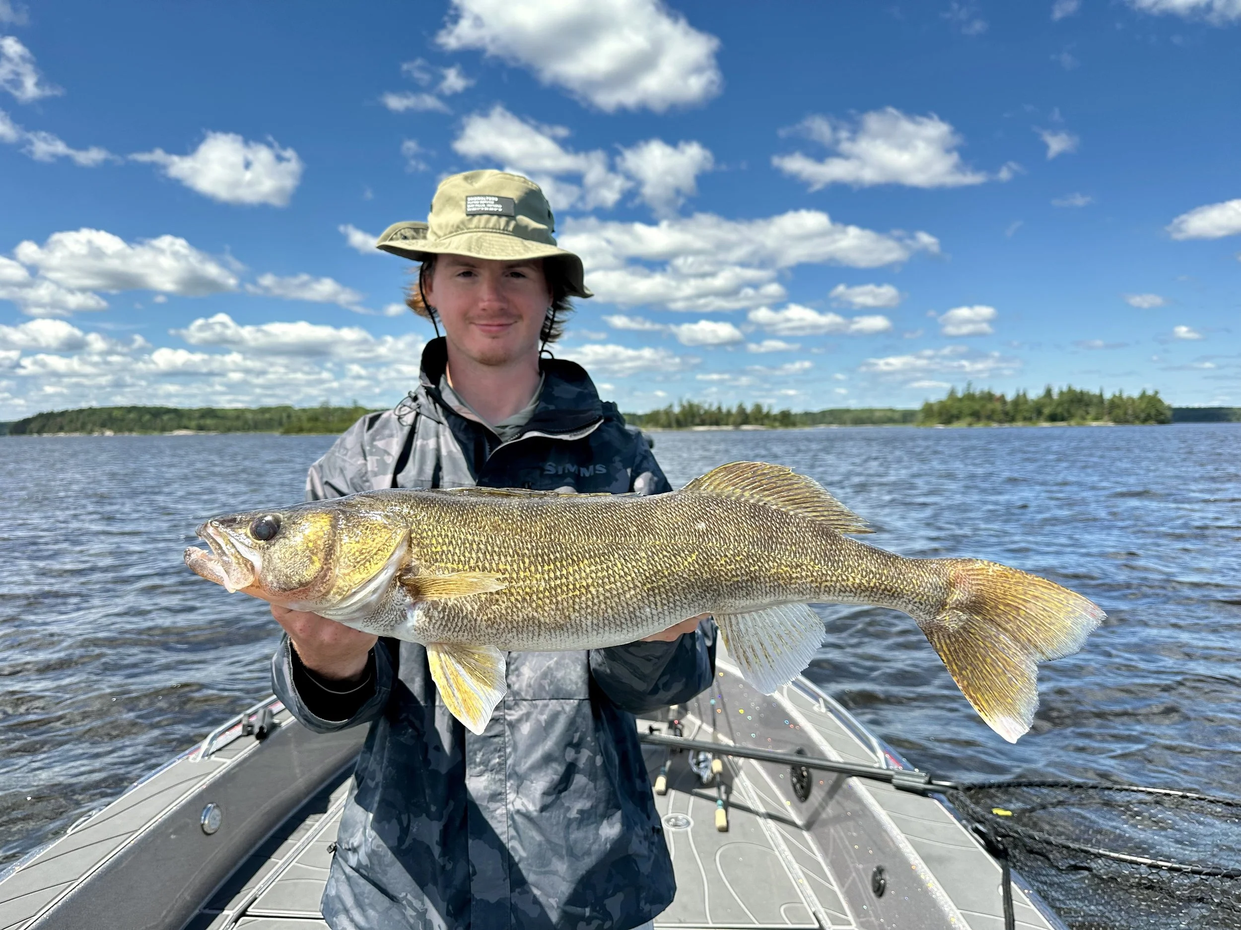 A young man in a fishing boat holding a large fish on a lake, with trees and a blue sky with clouds in the background.