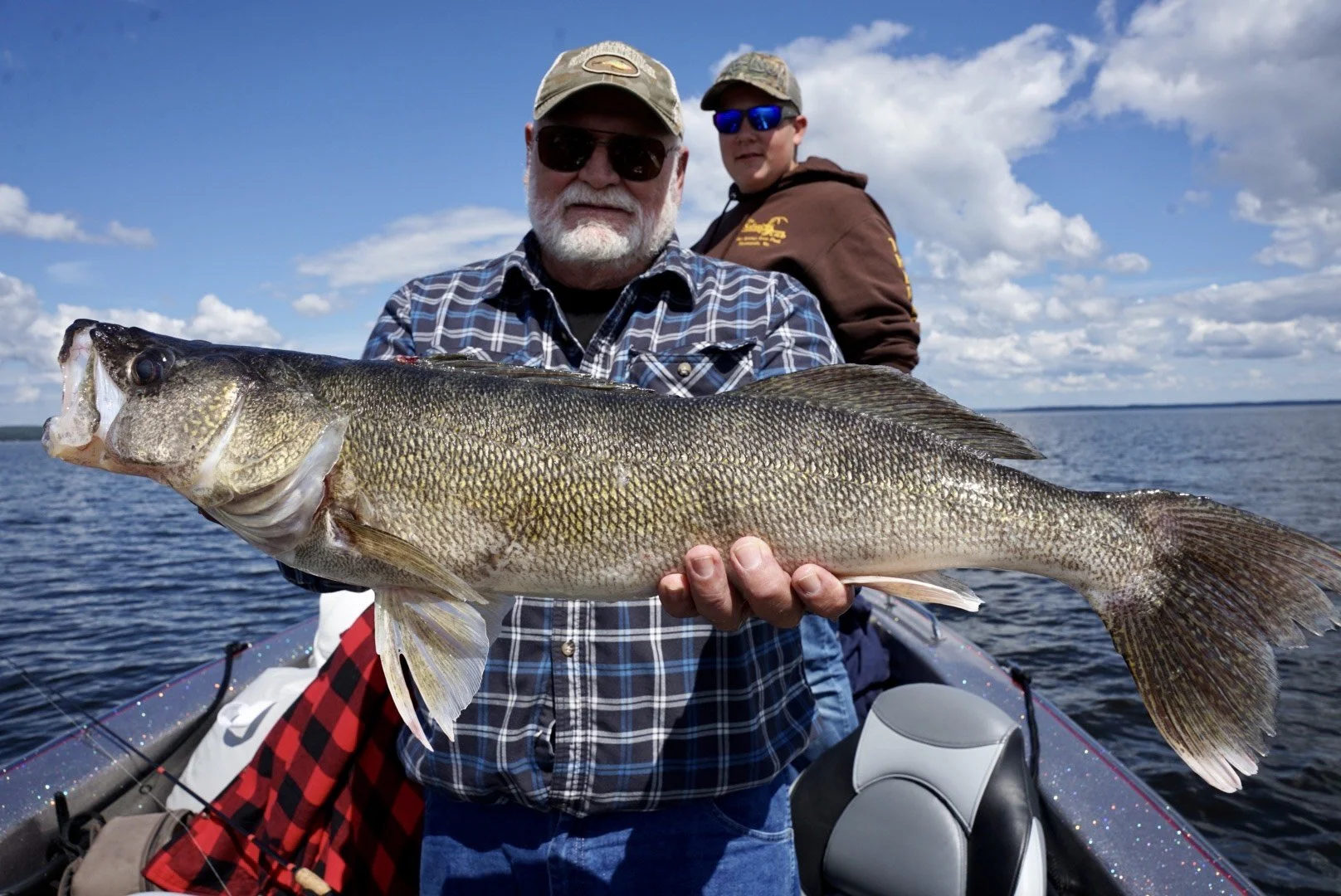 A man with sunglasses and a cap holding a large fish on a boat with a young boy standing behind him, both on a lake under a cloudy sky.