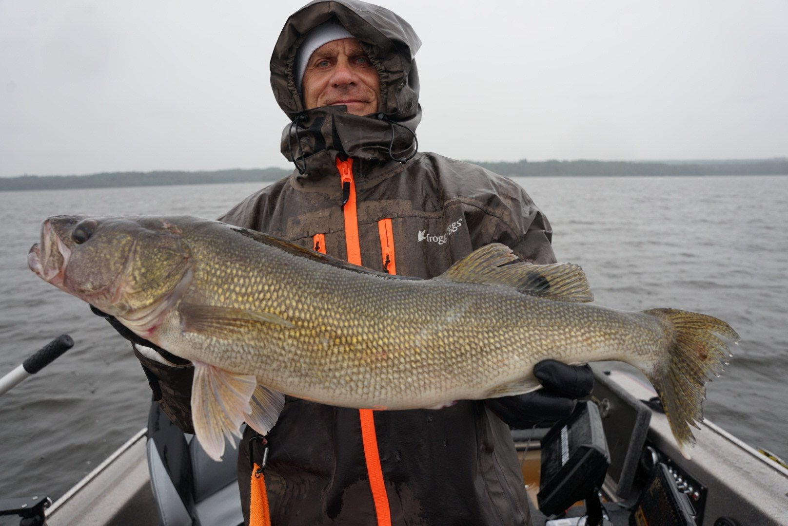 A man wearing a waterproof jacket and gloves holding a large fish on a boat in a body of water overcast sky.