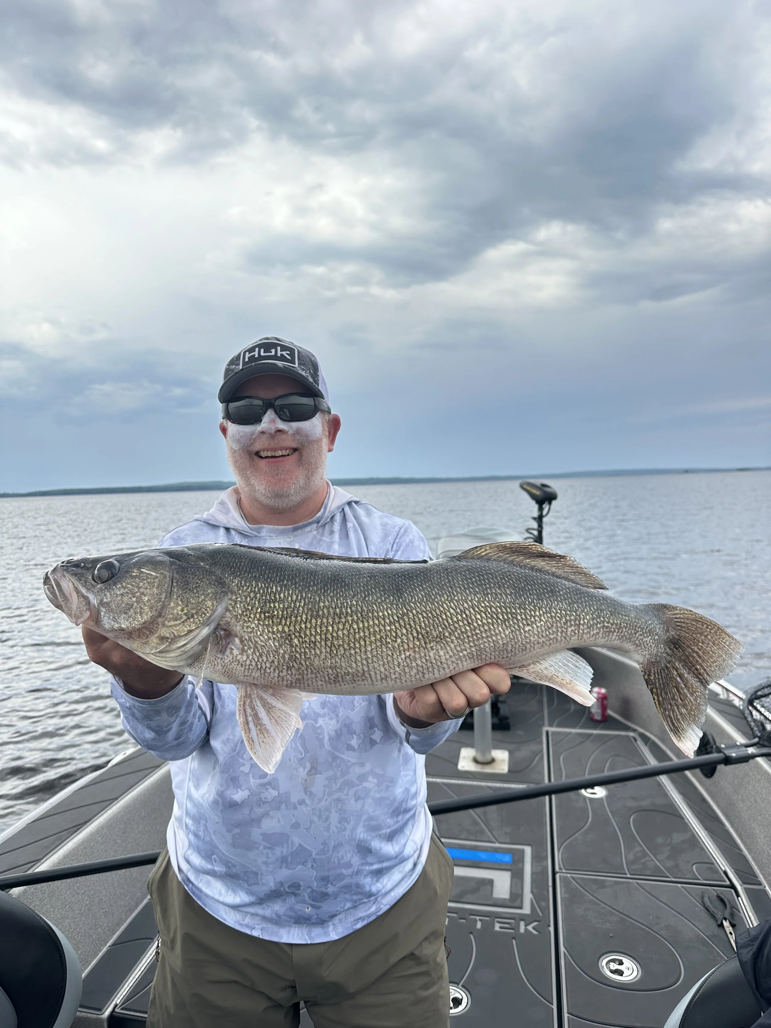 A man wearing sunglasses, a cap, and a hoodie is smiling and holding a large fish on a boat out on the water, with cloudy skies in the background.