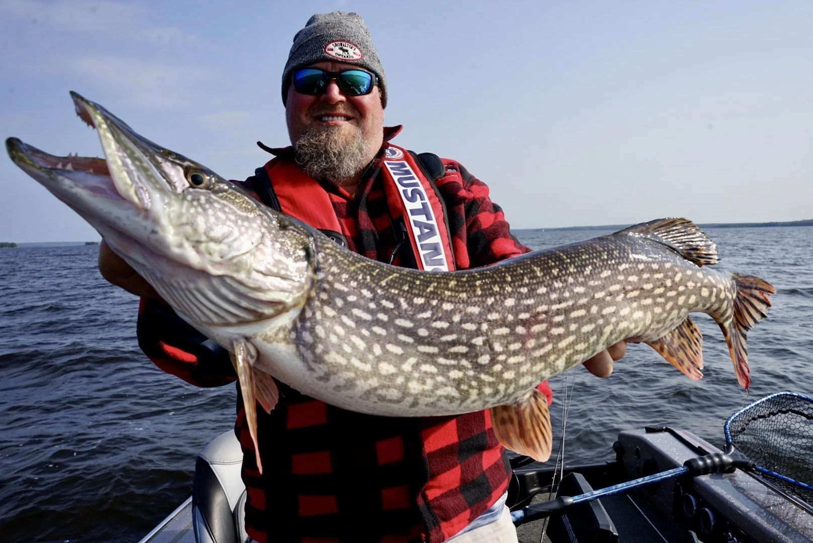 A man wearing sunglasses, a gray beanie, and a red flannel shirt holding a large fish on a boat out on the water, smiling at the camera.