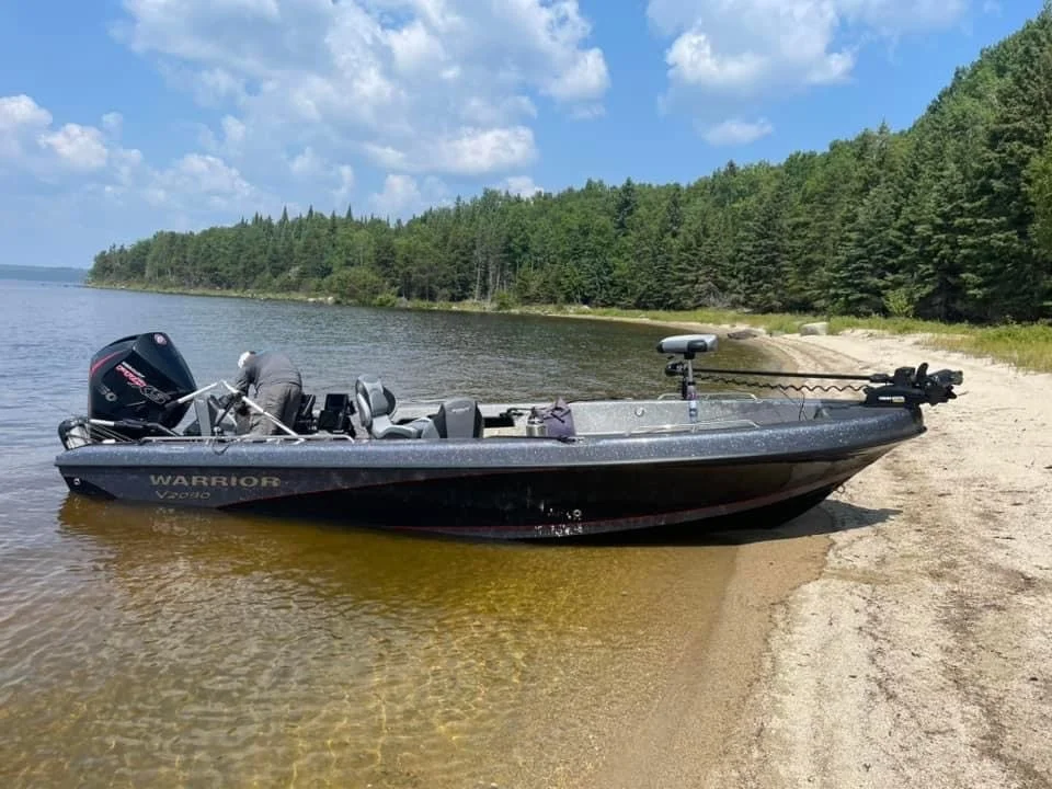 A black boat moored on a sandy shoreline with a forested area and blue sky in the background.