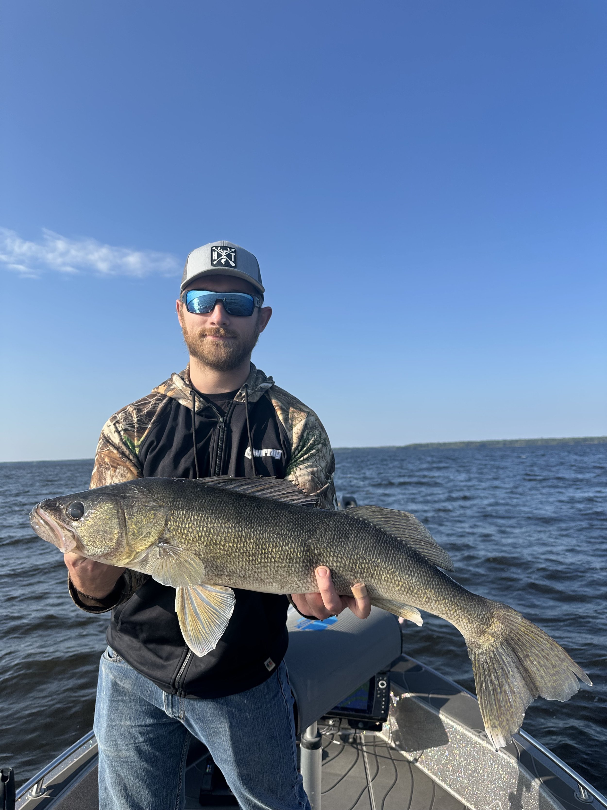 Man in camo jacket and sunglasses holding a large fish on a boat, with water and a blue sky in the background.