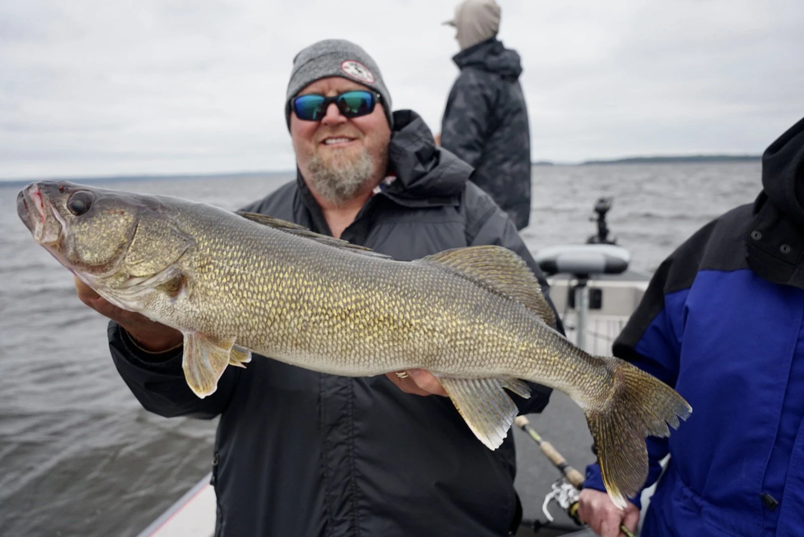 Man holding a large fish (walleye) on a boat with water and overcast sky in the background.