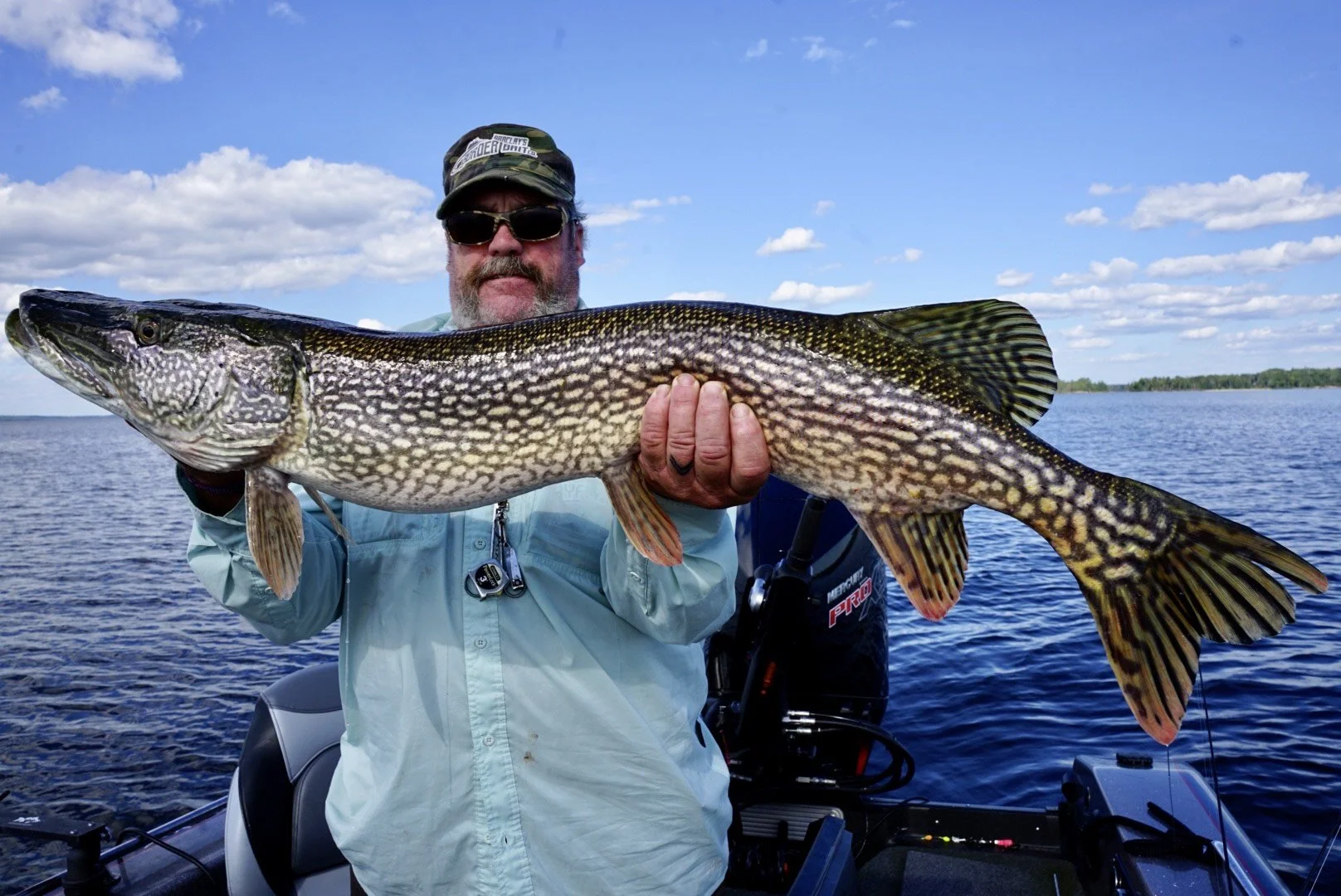 A man in sunglasses and a cap holding a large fish on a boat in a lake under a blue sky with clouds.
